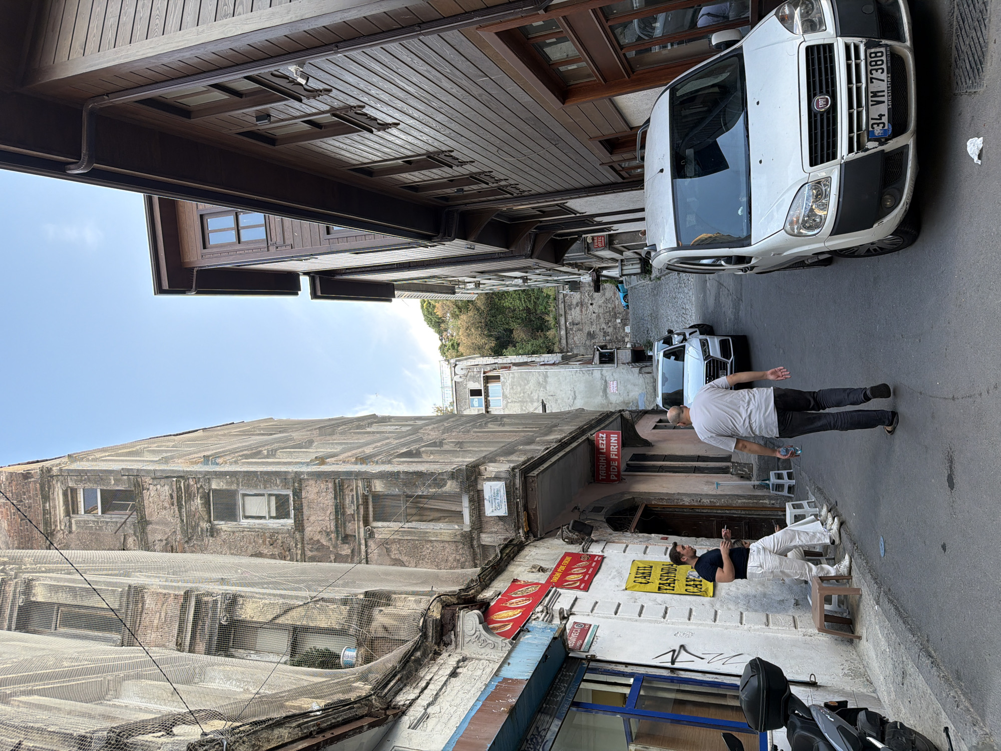 In a narrow, cobblestone street in Istanbul, Turkey, an aged, multi-story building covered in netting stands to the left, its facade weathered and showing signs of decay. Adjacent to it, a shop with a blue-framed window displays Turkish signage, with two men standing outside the shop; one in a black t-shirt and white pants is seated on a stool while smoking, and another in a dark t-shirt and white pants stands near him. On the right side of the street, a traditional wooden building with dark brown siding and multiple windows lines the road. A white Fiat minivan, with the license plate "34 VM 7388", is parked on the right side of the street, facing uphill. Further up the street, a white Audi SUV is visible. The sky above is clear blue, and a green hillside with trees is visible in the background at the top of the street. The overall scene depicts a typical street in a historical district of Istanbul.