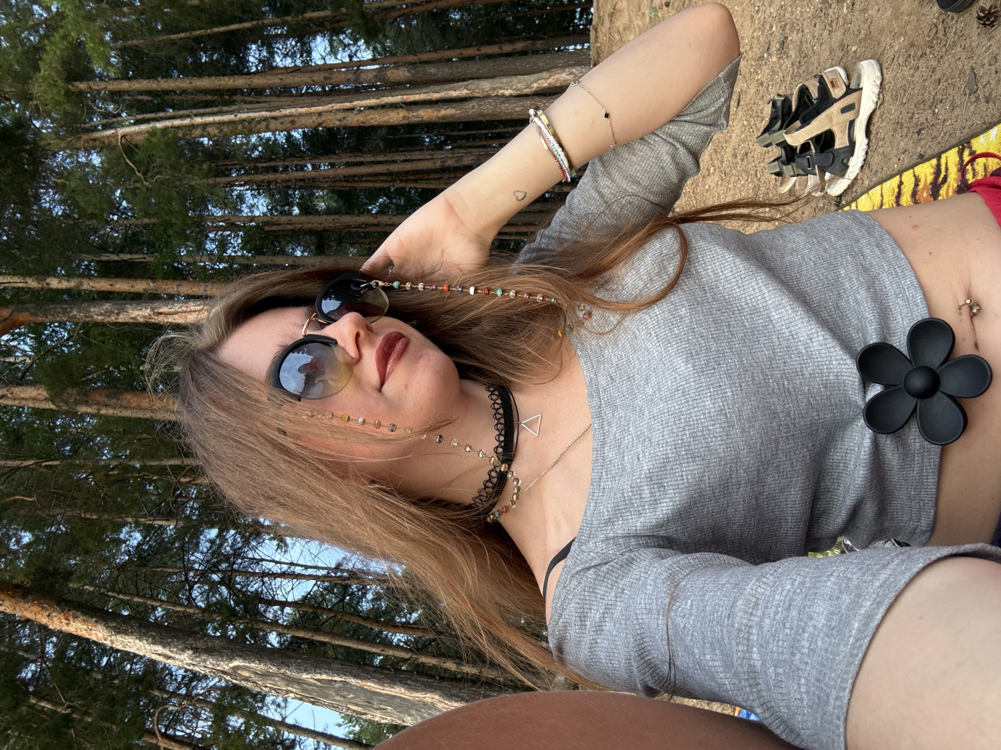 In an outdoor setting with a dense forest of tall pine trees in the background, a young woman with long auburn hair is captured in a selfie. She is wearing a grey off-the-shoulder top, sunglasses with a beaded chain, a black choker necklace with a triangular pendant, and multiple bracelets on her right arm. A black flower-shaped clip is attached to her top at the waist, and a belly button piercing is visible. A pair of beige and black sandals are placed on the ground behind her, along with a brightly colored mat. The lighting suggests it is daytime, possibly late afternoon due to the warm tones.