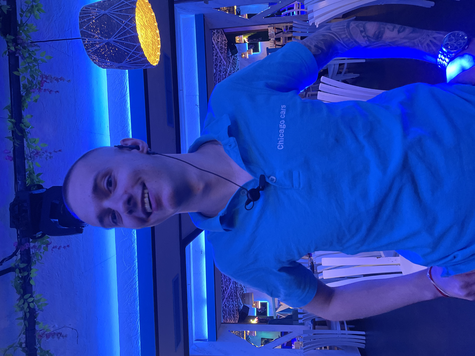 The image features a man smiling directly at the camera within a modern, illuminated bar or restaurant setting, likely at night in Plovdiv, Bulgaria. He has a shaved head and is wearing a bright blue polo shirt with the text "Chicago cars" printed vertically on the right side of his chest. He is also wearing a small black headset with a microphone attached near his collar. His left arm, visible on the right side of the frame, displays extensive tattoos and a luminous watch, while a thin red bracelet is visible on his right wrist.
The environment is characterized by prominent blue LED strip lighting running along walls and structural elements, casting a cool, ambient glow across the scene. Contrasting this blue light, a large, decorative woven lamp with a warm, yellowish glow hangs in the upper left portion of the image. To the left, some artificial foliage with small purple flowers adds a touch of natural decor. In the background, white chairs and tables suggest a dining or lounge area, along with decorative wall panels, indicating a contemporary entertainment venue. The man's cheerful expression contributes to the lively and inviting atmosphere of the scene.