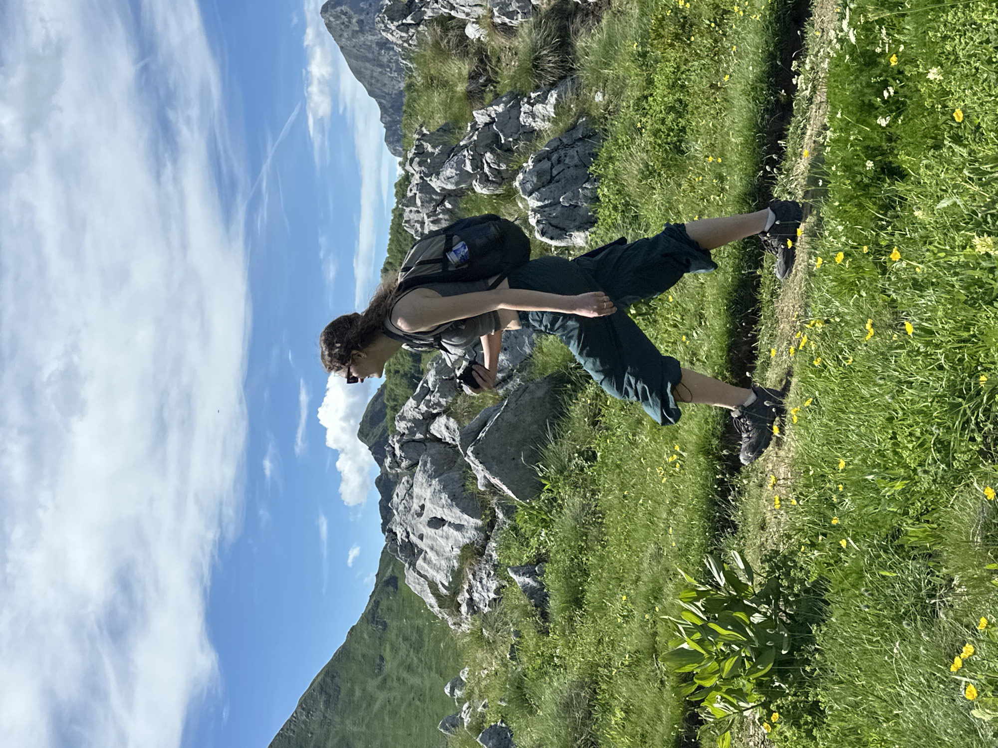 A young woman with curly brown hair and sunglasses is hiking on a rocky, grassy mountain trail. She is wearing a dark gray crop top, dark green cargo pants, and black hiking boots. She has a black backpack on, and is holding a black camera in her right hand. The woman is walking from left to right, in profile. She is on a narrow dirt path that winds through a landscape of green grass, scattered yellow wildflowers, and large grey rocks. In the background, there are more rocky mountains covered in green vegetation, with a bright blue sky dotted with white clouds above. The scene is outdoors and appears to be in a natural, mountainous environment, possibly in or near the city of Dolja, Montenegro.