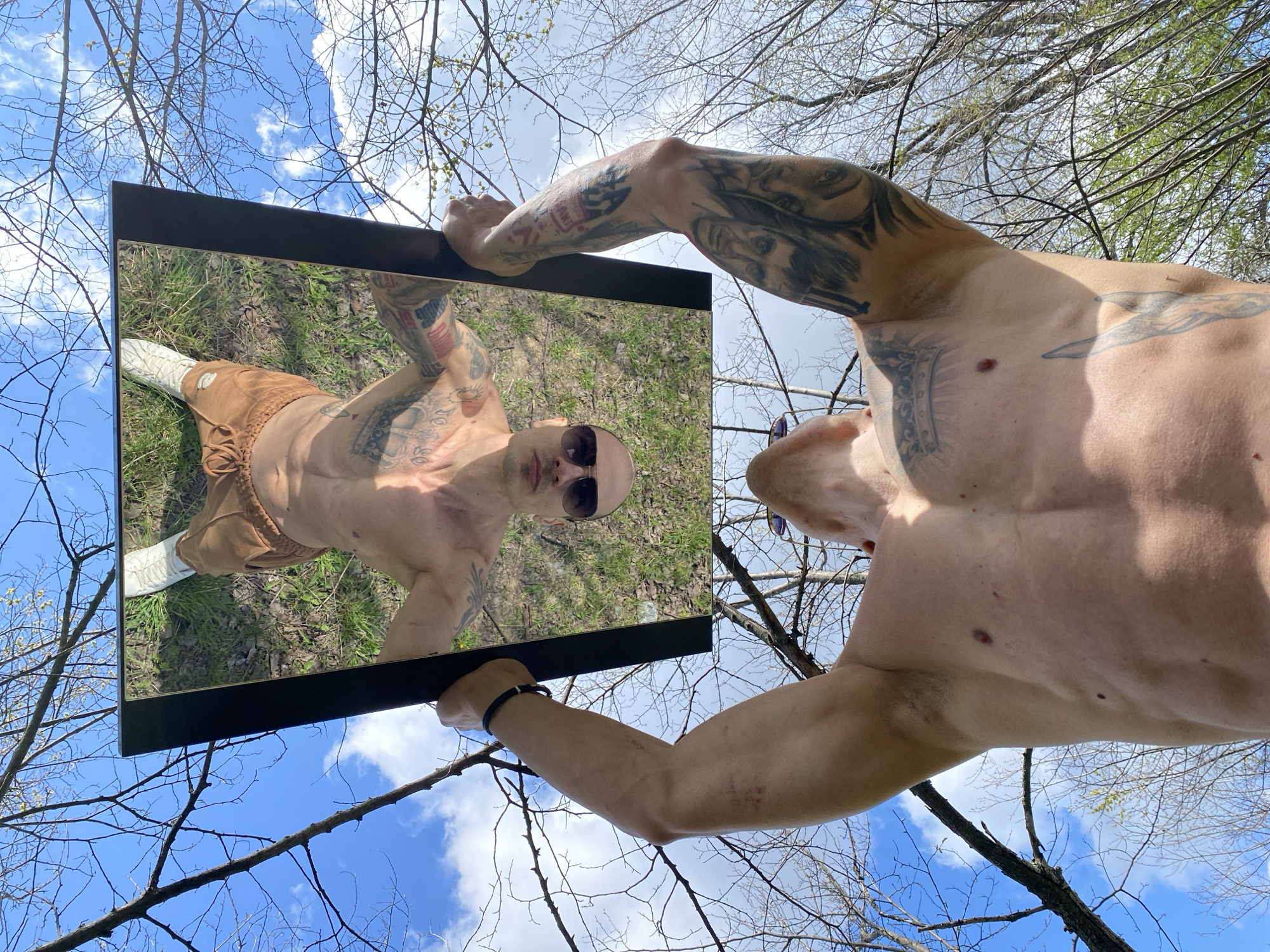 A shirtless, muscular man with numerous tattoos is holding a rectangular mirror in an outdoor setting, reflecting his image. He is wearing sunglasses and brown pants with white shoes in the reflection. The background consists of trees with bare branches against a blue sky with scattered clouds. The location is not evident and cannot be determined to be in Pleven, Bulgaria, from the provided image.