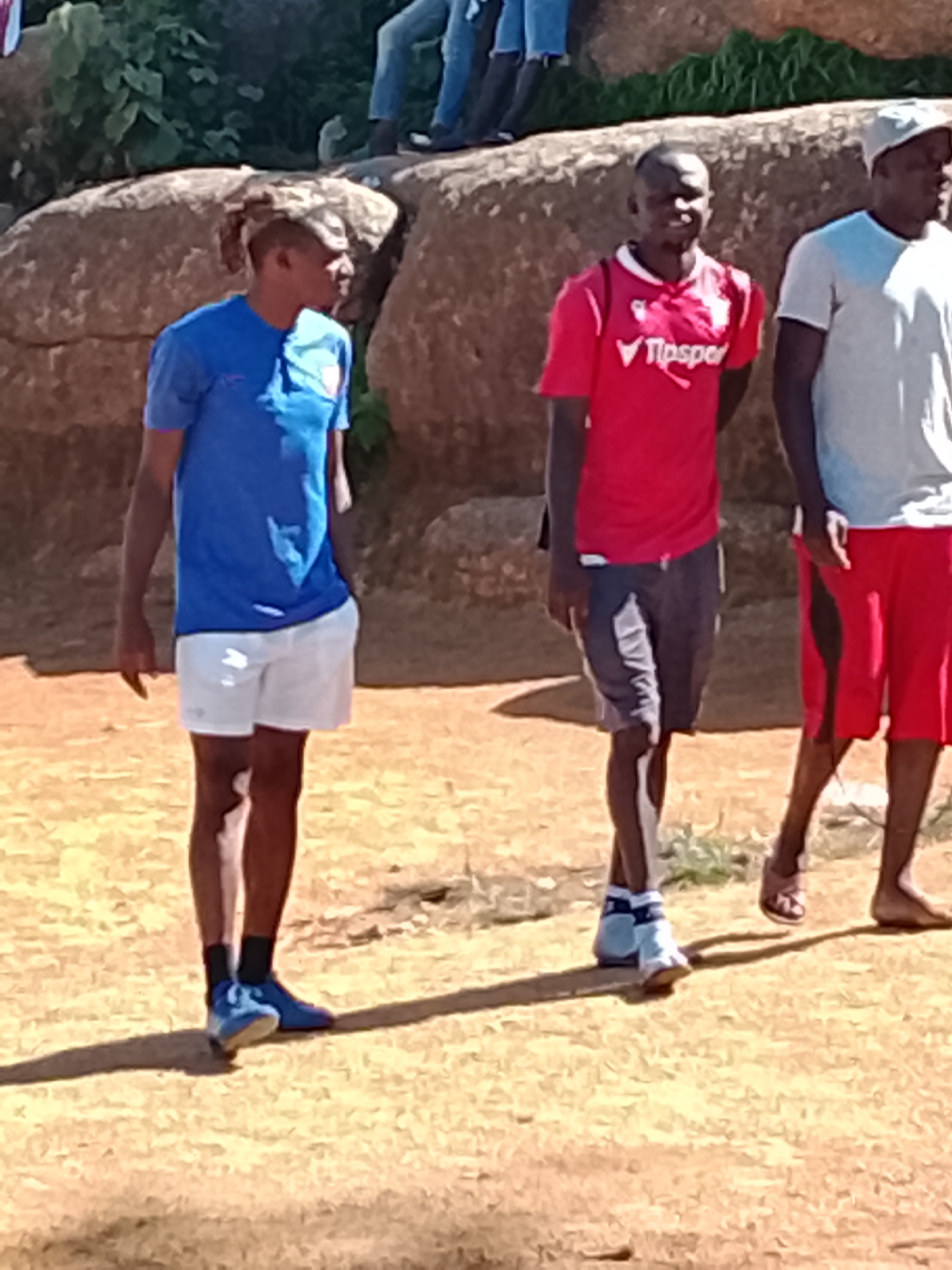 In Jos, Nigeria, three men stand outdoors on a sunlit dirt ground. The man on the left wears a blue t-shirt and white shorts. The man in the center sports a red t-shirt with a white logo and grey shorts. The man on the right is clad in a light-colored t-shirt and red shorts. Behind them, large, beige rocks form a natural backdrop, with green foliage visible above. The scene appears to be an informal gathering or activity.