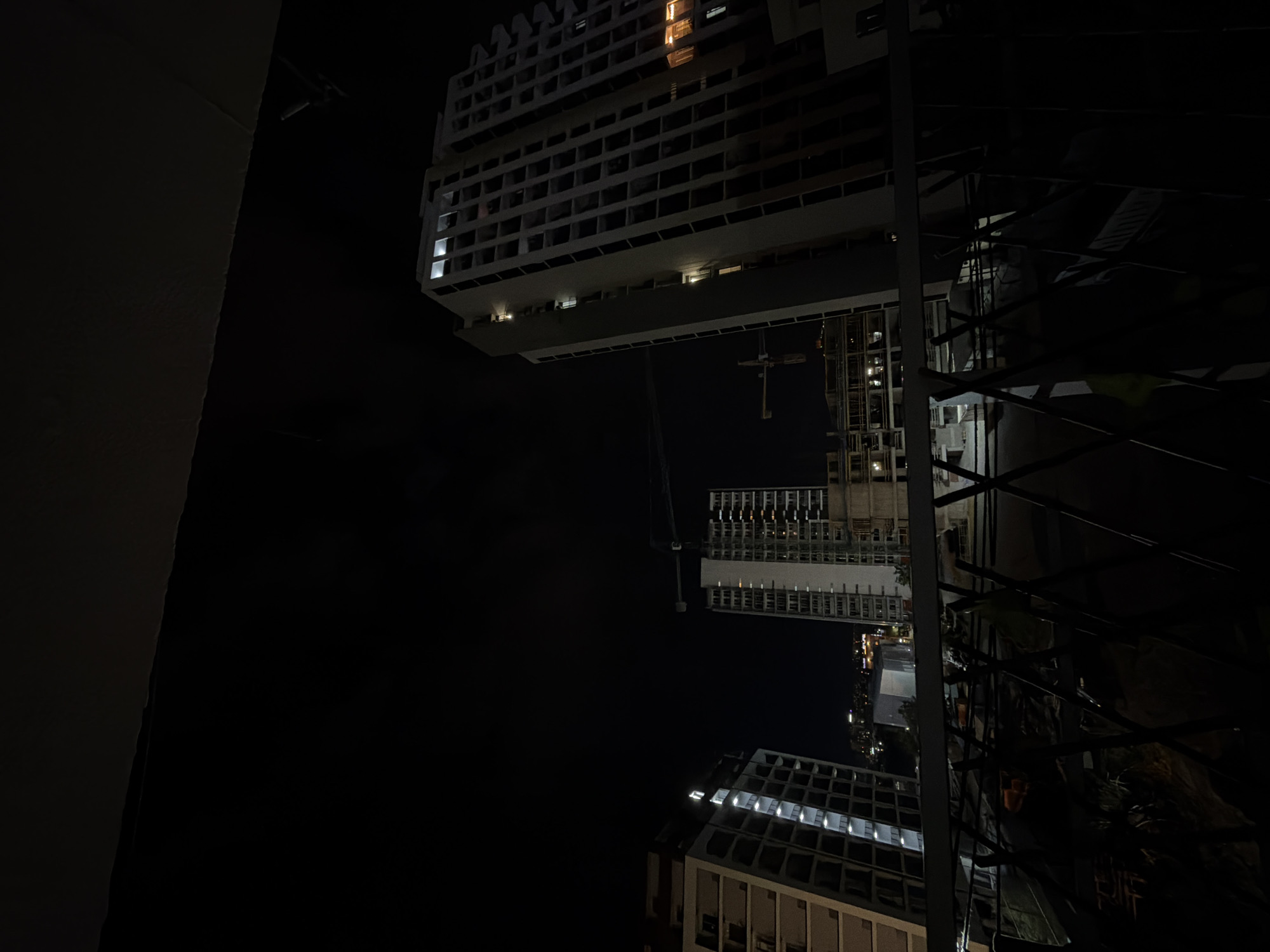 The image captures a nighttime view of a city skyline, likely in Tbilisi, Georgia, featuring several unfinished high-rise buildings. The scene is dominated by darkness, with the illuminated windows of the buildings providing the primary light source. In the foreground, a dark silhouette of a railing or fence is visible, suggesting the photograph was taken from a balcony or elevated vantage point.
Two prominent buildings are centrally positioned. The building on the right is a tall, modern structure, its facade a grid of balconies or open window frames. Some windows are lit, casting a warm glow, while others are dark, indicating ongoing construction. A construction crane is visible to the left of this building, its boom extending upwards into the night sky.
To the left of the central buildings, another high-rise structure is depicted. This building also features a grid-like design with illuminated windows. Its silhouette is slightly less defined than the central building on the right, suggesting it may be further away or also under construction.
In the background, a cluster of lower buildings with distant lights can be seen, hinting at a more developed part of the city. The overall atmosphere is one of urban development and construction, captured during the nocturnal hours. There are no people visible in the image. The environment appears to be an urban setting with significant construction activity.
