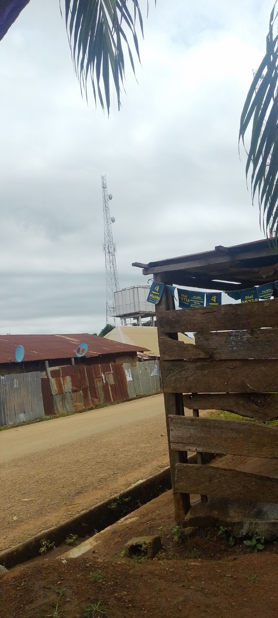 The image captures a view of a street in Baissa, Nigeria, under an overcast sky. In the foreground, a rustic wooden fence made of rough-hewn planks partially obscures the scene. To the left of the fence, a dirt road runs alongside a drainage ditch. Further down the road, a row of buildings with corrugated metal roofs is visible. One building has a rusted metal roof and a weathered metal siding. Two satellite dishes are mounted on this building's roof. In the background, a tall telecommunications tower stands against the cloudy sky, accompanied by a water tank structure. Several small, triangular flags advertising a service called "MoMo" and "Pay Bills" are strung along the top of the wooden fence. The overall impression is one of a rural or semi-urban environment with basic infrastructure.