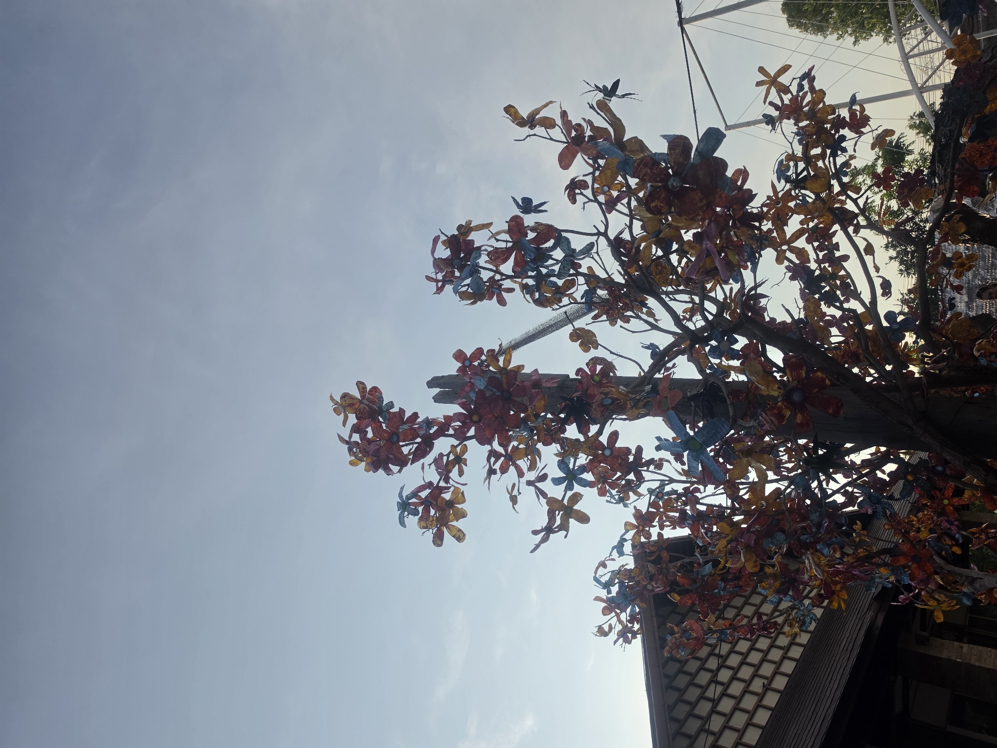 The image presents an upward perspective of a vibrant outdoor art installation against a clear, bright blue sky with faint, wispy clouds, indicating fair daytime weather in Pattaya City, Thailand. The primary subject is a tree-like structure, possibly an actual tree or an artificial sculpture, adorned with numerous colorful artificial flowers and several small birds. These decorative elements appear to be crafted from recycled plastic materials, likely plastic bottles, showcasing a creative use of sustainable resources.
The artificial flowers are painted in a cheerful palette of red, orange, yellow, blue, and purple, creating a striking contrast against the pale sky. Several artificial birds, also brightly colored and made from similar materials, are perched among the branches, adding to the whimsical nature of the display. In the lower right portion of the frame, part of a building's dark-colored roof is visible, featuring a distinct patterned or tiled section. Subtle structural elements, such as wires or poles, can be faintly discerned against the sky in the upper sections, suggesting a larger surrounding structure or an intricate mounting for the art piece. No specific activity or event is taking place, as the scene is a static decorative display. There are no people visible, and no discernible text in the image. The overall impression is one of artistic ingenuity and a colorful celebration of recycled materials in a public setting.