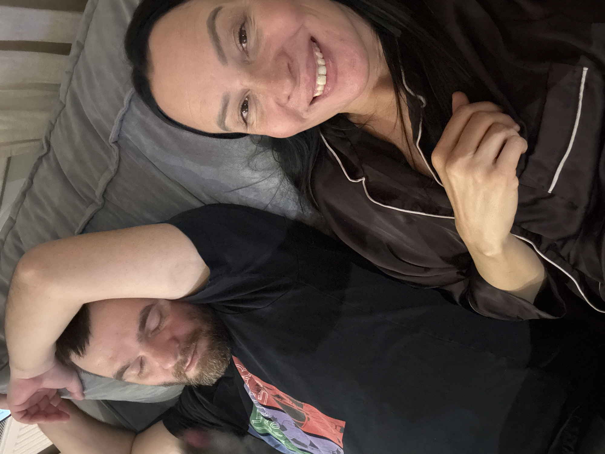 A woman with long, dark hair and wearing a dark satin pajama top is smiling at the camera. She is positioned above a man who is lying on his back with his eyes closed. The man has a beard and is wearing a black t-shirt. They are both lying on a gray pillow or bedding. The image appears to be taken in a bedroom setting. There is no information to suggest this image was taken in the city of Chaikovsky, Russia.
