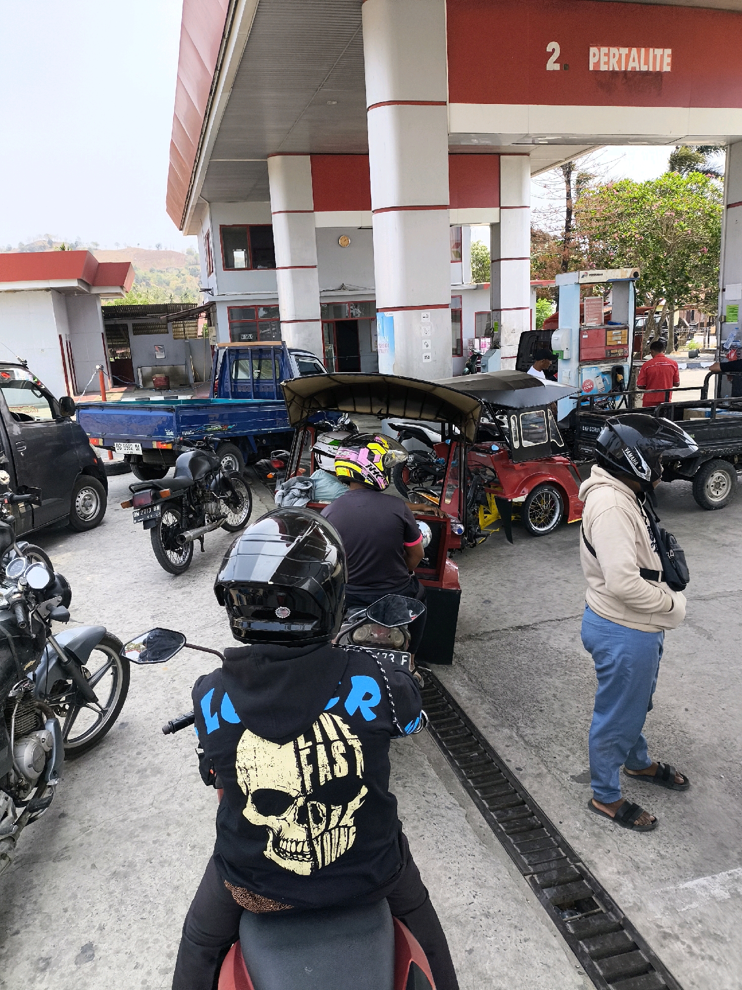 The image depicts a busy outdoor scene at a Pertamina gas station in Indonesia. In the foreground, a person wearing a black hoodie with a skull graphic and a black helmet is seated on a red motorcycle. To their right, another person in a tan hoodie and blue pants stands, looking towards the gas pumps.
Several vehicles are present, including a black car on the left, a blue pickup truck in the middle, and a red motorized three-wheeled vehicle with a canopy. Motorcycles are parked or waiting in line. Two men in red shirts appear to be gas station attendants, interacting with customers or managing operations.
The gas station features red and white structural elements, with signs indicating "PERTAMINA" and fuel types. The environment appears to be a relatively flat, paved area with some vegetation and a hilly landscape in the background under an overcast sky. The overall impression is one of a typical, functional gas station serving a variety of vehicles.