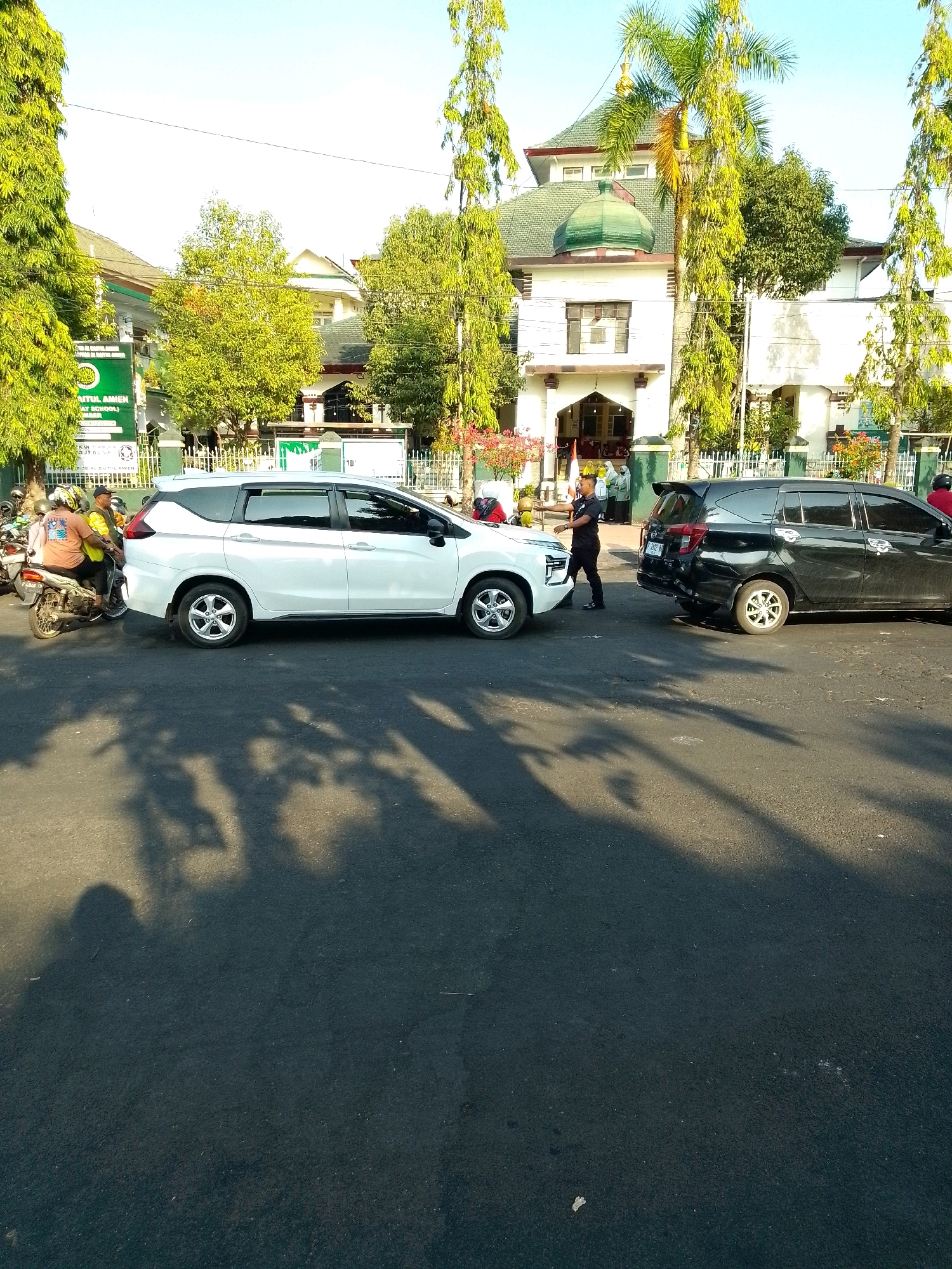 The image depicts a street scene in Jember, Indonesia, in front of a building with traditional Indonesian architectural elements. A white SUV is parked on the right side of the road, and a black SUV is parked behind it, also on the right. A man in a black uniform, possibly a security guard, is standing between the two SUVs, facing the white SUV. On the left side of the road, a motorcycle with two people on it is parked. The background shows a building with a green domed roof, reminiscent of a mosque or a traditional Indonesian house. Trees line the street, casting shadows on the asphalt. There are also people visible in the doorway of the building. The sky is clear and blue, suggesting it is daytime. The overall impression is of a busy street with vehicular and pedestrian activity.