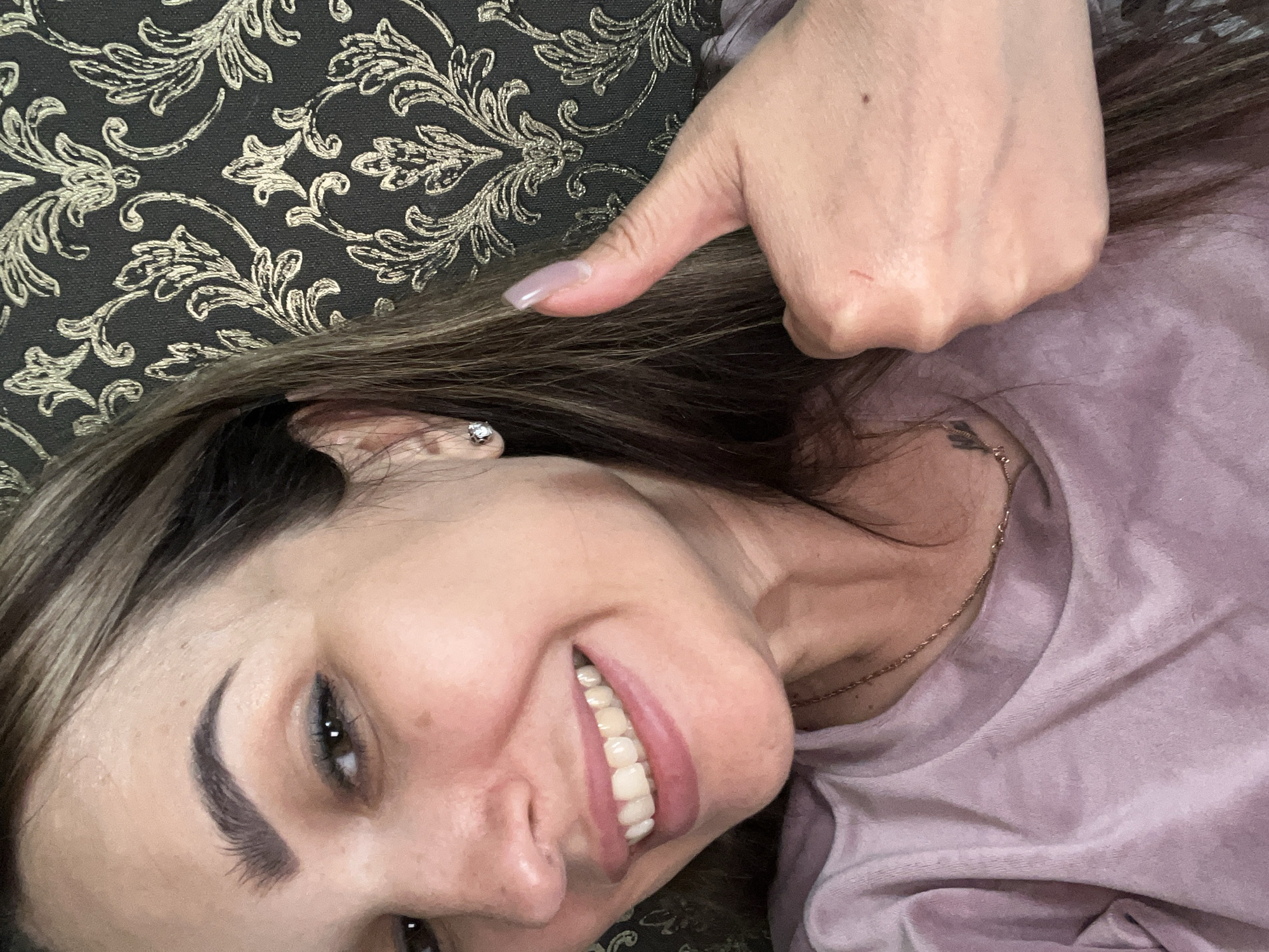 A woman with dark brown hair and brown eyes smiles and gives a thumbs-up. She is wearing a pink t-shirt and a gold necklace. She has on a diamond stud earring and her fingernails are light pink. The background is a dark wallpaper with a gold floral pattern. The image is a close-up of the woman's face and hand.