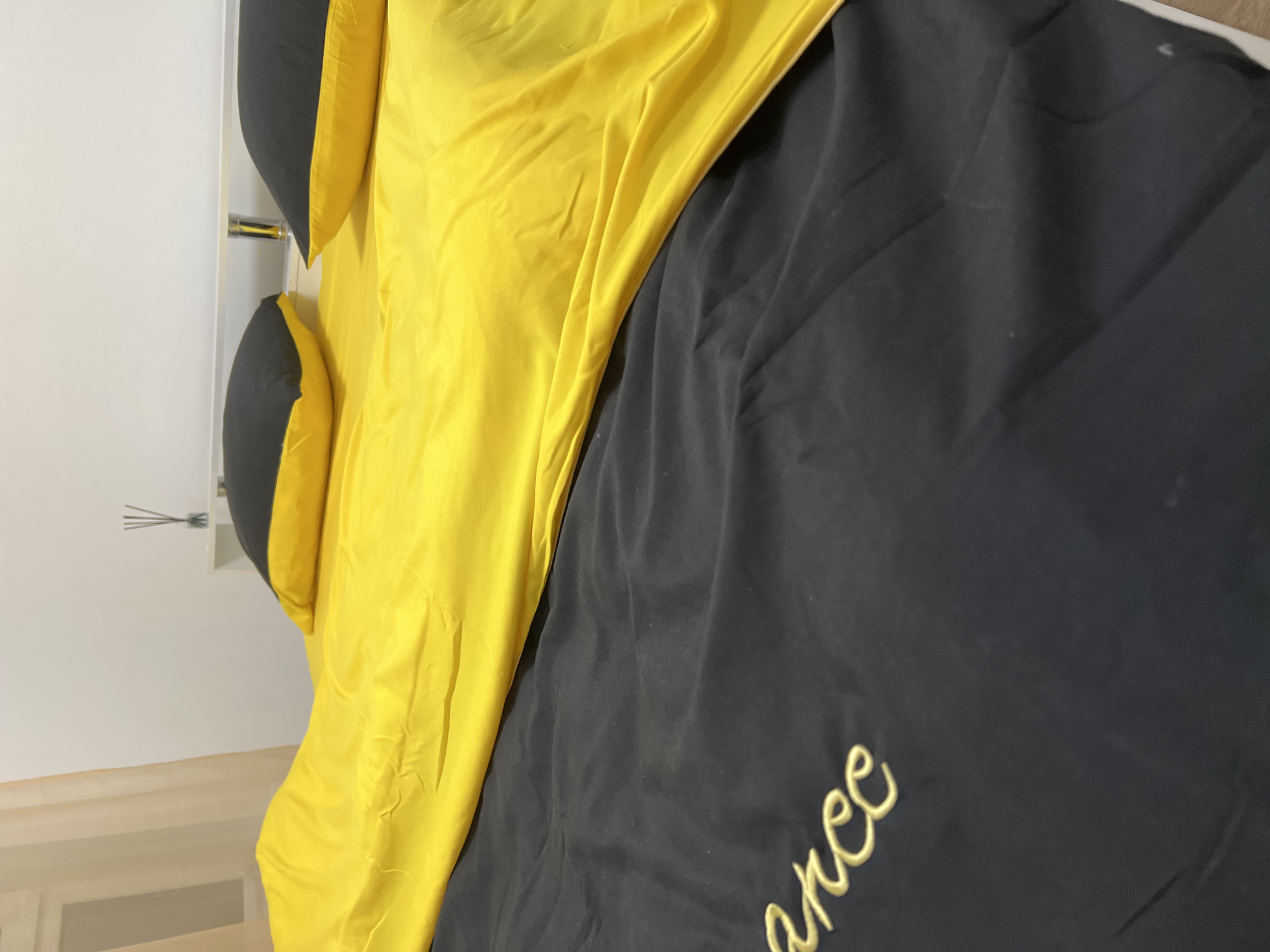 The image shows a close-up view of a bed with yellow and black bedding. Two black pillows with yellow trim are visible on the yellow comforter. The bedding appears to be made of a smooth, synthetic material. In the background, a white headboard and a white wall are visible. A sheer beige curtain hangs to the left of the frame, and a small vase with reeds is on a shelf behind the pillows. The lower portion of the image shows the black duvet cover with gold embroidery that reads "arc". The image does not provide any information about the location in the city of Yekaterinburg, Russia.