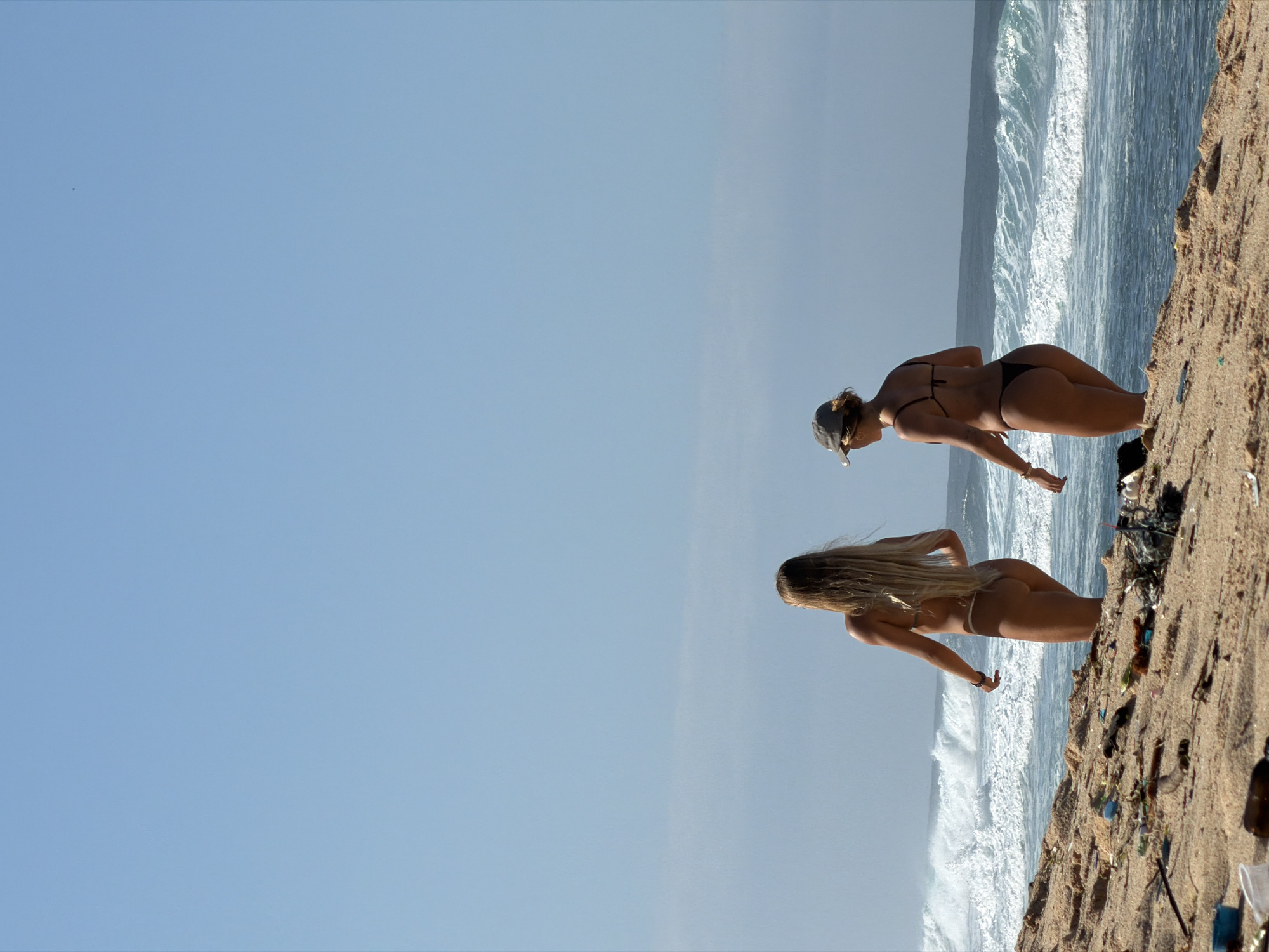 Two women in bikinis stand on a sandy shore beside an ocean with breaking waves under a clear blue sky. The shore appears to be cluttered with debris. The image appears to have been taken at Padang Padang Beach in Kuta Selatan, Indonesia.