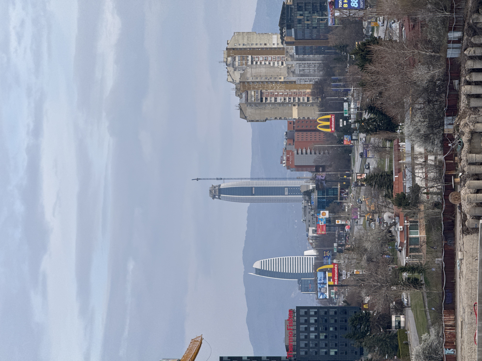 The image presents an urban scene in Sofia, Bulgaria. The foreground shows a concrete structure, leading to a street with light vehicular traffic. Billboards and signs are visible along the road. Several buildings dominate the skyline, with a prominent, modern skyscraper under construction. Behind the city, mountains are faintly visible through the haze. McDonald's restaurant is visible along with other businesses. The sky is overcast with gray clouds.