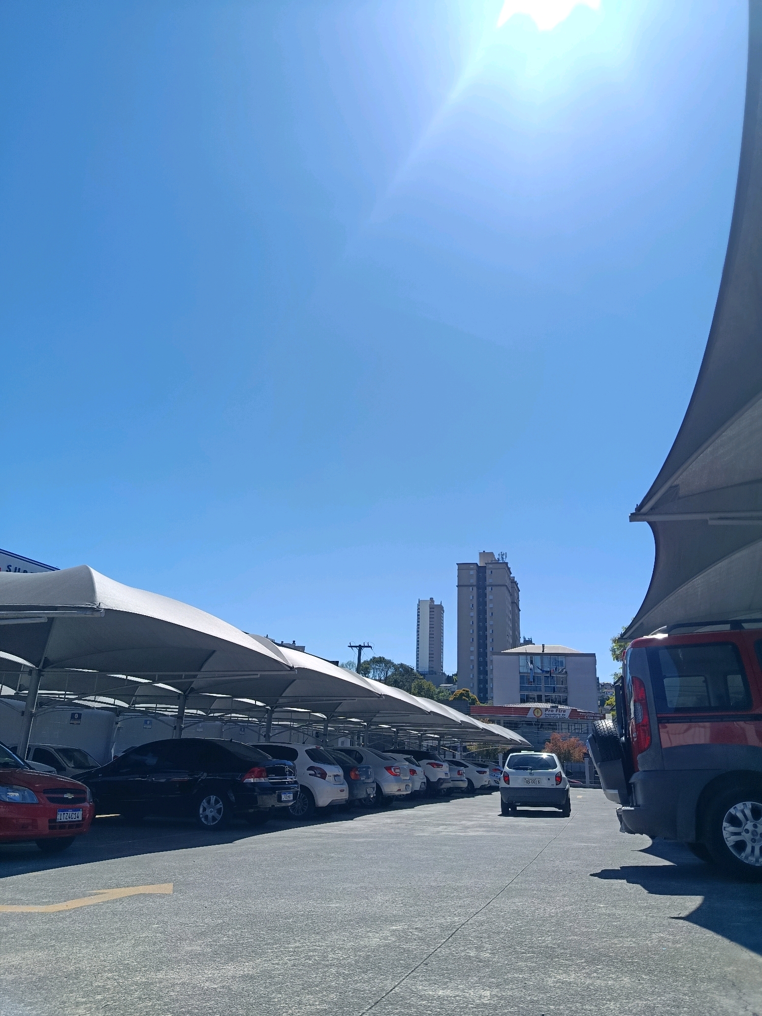 The image captures a street-level view of a parking lot under a bright, clear blue sky. The parking lot is covered by a series of white fabric canopies supported by metal poles, providing shade for the parked vehicles. Several cars are visible in the lot, including a red car in the foreground on the left, a black sedan, a white hatchback, and a red van or SUV on the right. The ground is paved asphalt, marked with yellow lines, including a directional arrow.
In the background, tall buildings rise into the sky, suggesting an urban setting. One prominent building appears to be a skyscraper, with several other smaller buildings visible between it and the foreground. Green trees can be seen scattered amongst the buildings. The overall scene is of a well-lit, outdoor parking area in a city. The license plates visible on the cars suggest they are from Brazil, with one license plate reading "IT Z4C14" and another "TOS 6G19".
Based on the license plate format and the architectural style of the buildings, this scene is likely located in Caxias do Sul, Brazil. The sun is high in the sky, indicated by the strong glare and shadows, suggesting it is midday. There are no people visible in the image. The atmosphere is one of a typical urban environment on a sunny day.