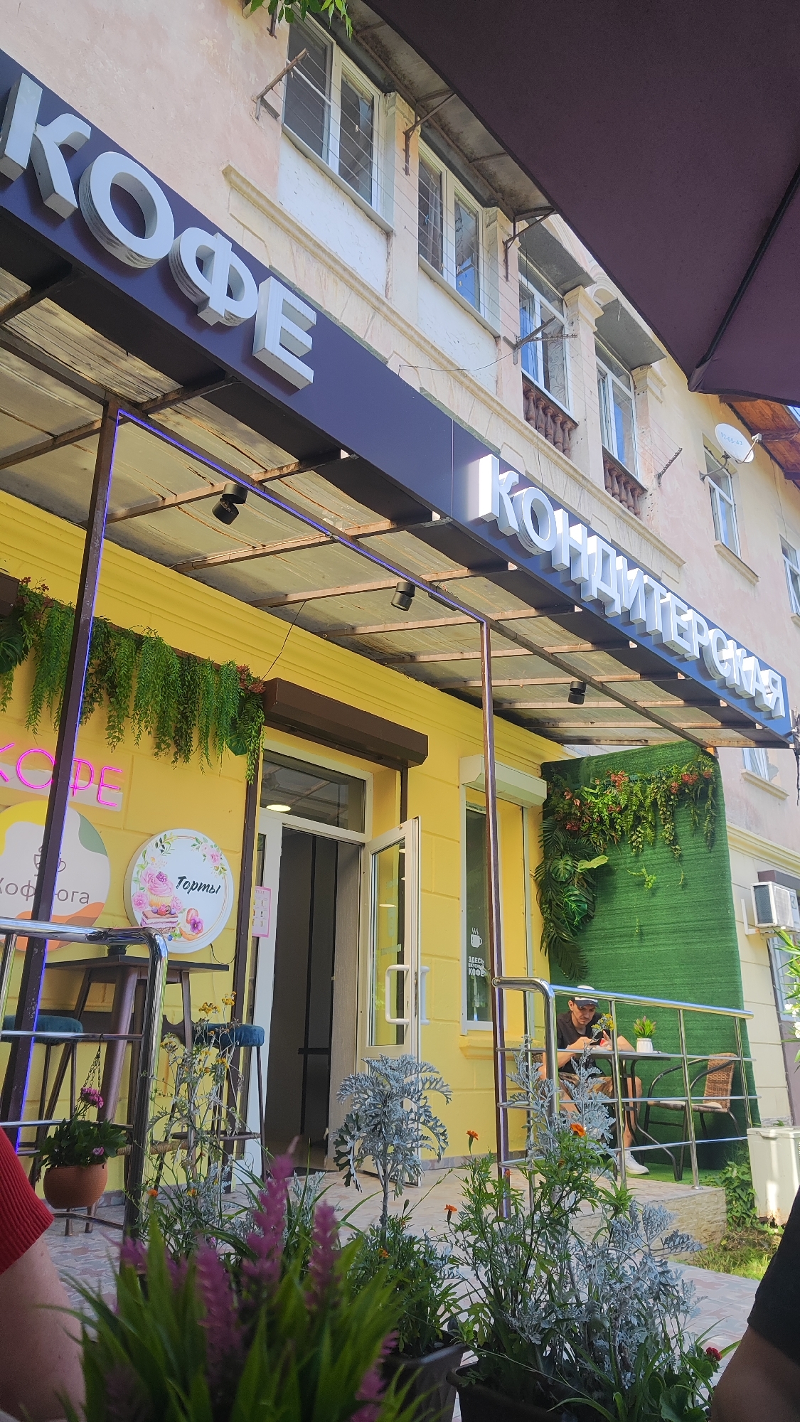 The image shows the exterior of a cafe with a bright yellow facade. A large sign above the entrance reads "КОФЕ" in white, three-dimensional letters on a dark blue background. To the right, another sign says "КОНДИТЕРСКАЯ" in similar white lettering.
There are several round signs on the yellow wall. One, illuminated in pink neon, says "КОФЕ". Another, with a floral design, has the word "Торты" (Cakes) written on it.
In front of the cafe, a covered outdoor seating area is visible. Potted plants with green and purple foliage are placed in the foreground. A person is sitting at a small table on a patio to the right, seemingly using their phone. The area appears to be well-lit, suggesting it is daytime, and the presence of plants and outdoor seating implies pleasant weather.
The setting appears to be a street-level establishment in a town or city, possibly in Dagomys, Russia, given the context. The overall impression is of a welcoming cafe with a focus on coffee and pastries.