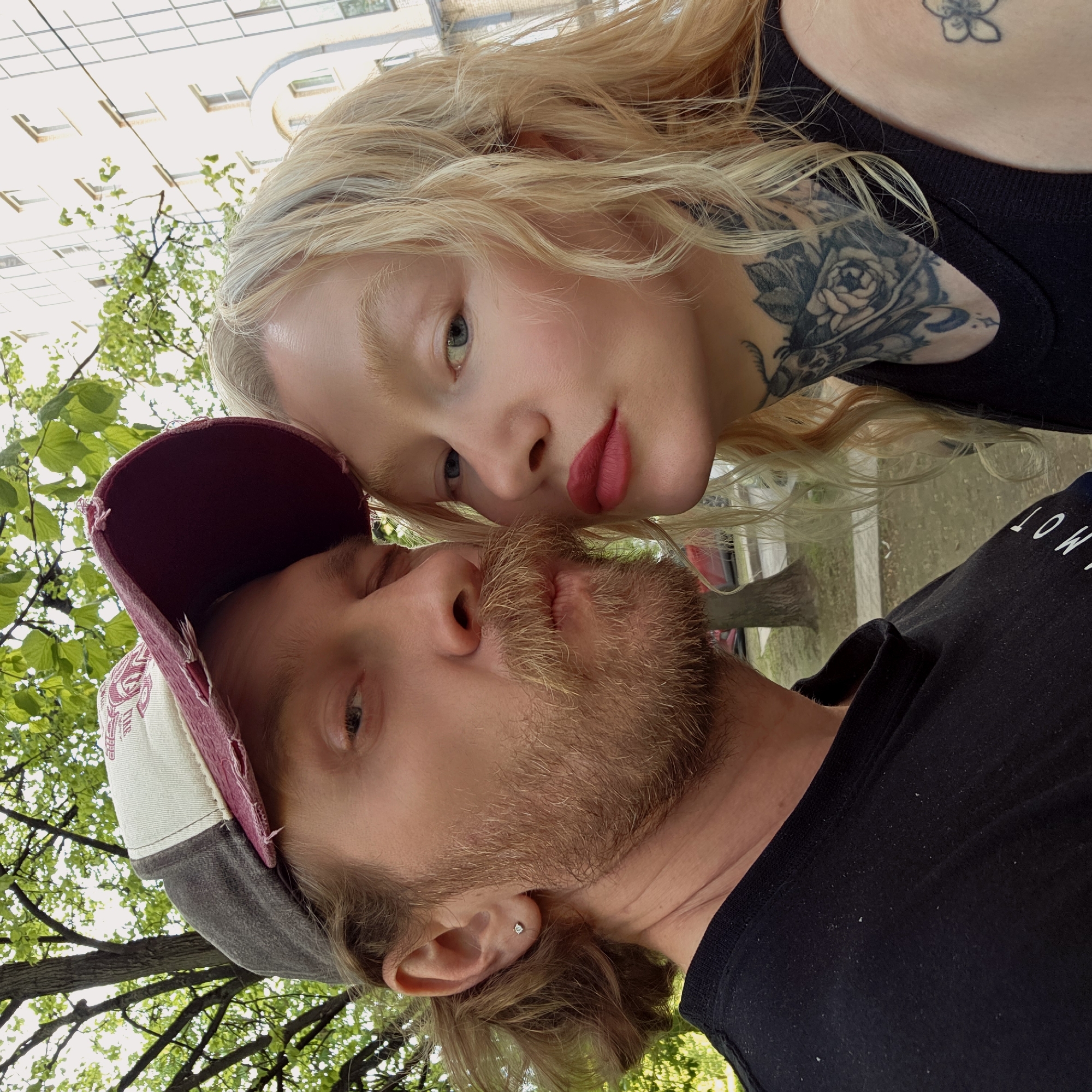 A blonde woman with fair skin, distinctive red lipstick, and neck tattoos and a fair-skinned man with a beard and cap pose closely together, presumably for a selfie. The background includes a tree with green leaves and a tall, multi-story building, which suggests an urban environment. Based on the architectural style of the building, it may be in Saint Petersburg, Russia.