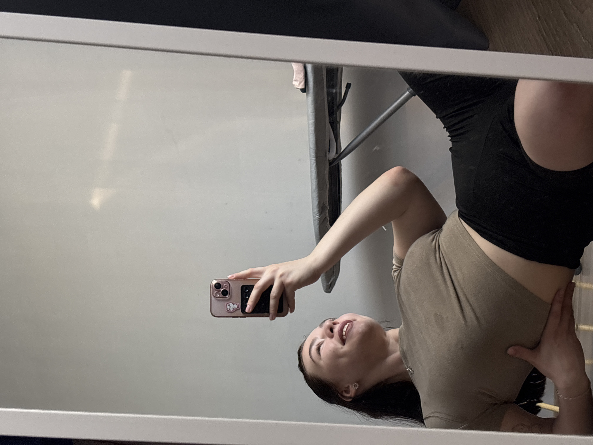 A woman is taking a selfie in a mirror. She is wearing a tan shirt and black shorts. The mirror is framed with a white border. In the background, there is a wall and an ironing board. The location is not identifiable as Perm, Russia.