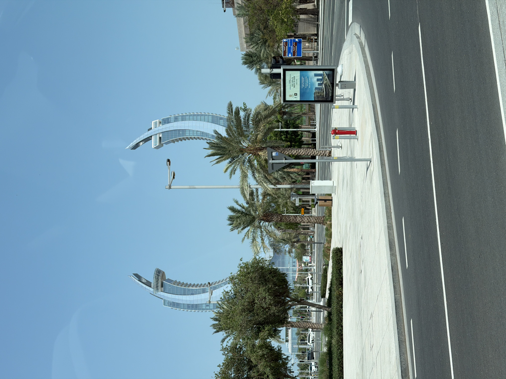 In a city scene under a clear blue sky, two distinctive curved skyscrapers with glassy exteriors rise in the background. In the mid-ground, a paved area features several palm trees and other greenery, with a street and sidewalk in the foreground. A bus stop shelter with a digital advertisement stands on the right side of the image, near a red fire hydrant. Traffic lights and street lamps are visible, along with several white vehicles on the road. The overall environment suggests a modern, urban setting with a focus on architecture and infrastructure.