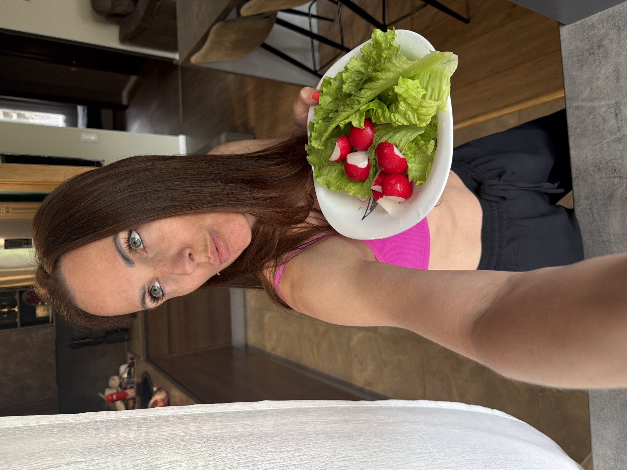 A woman with long brown hair and blue eyes is in a kitchen. She is wearing a pink sports bra and black drawstring pants. She is holding a white plate with lettuce and radishes. The kitchen has wooden cabinets and a tiled floor. The background shows a dining area with a table and chairs.