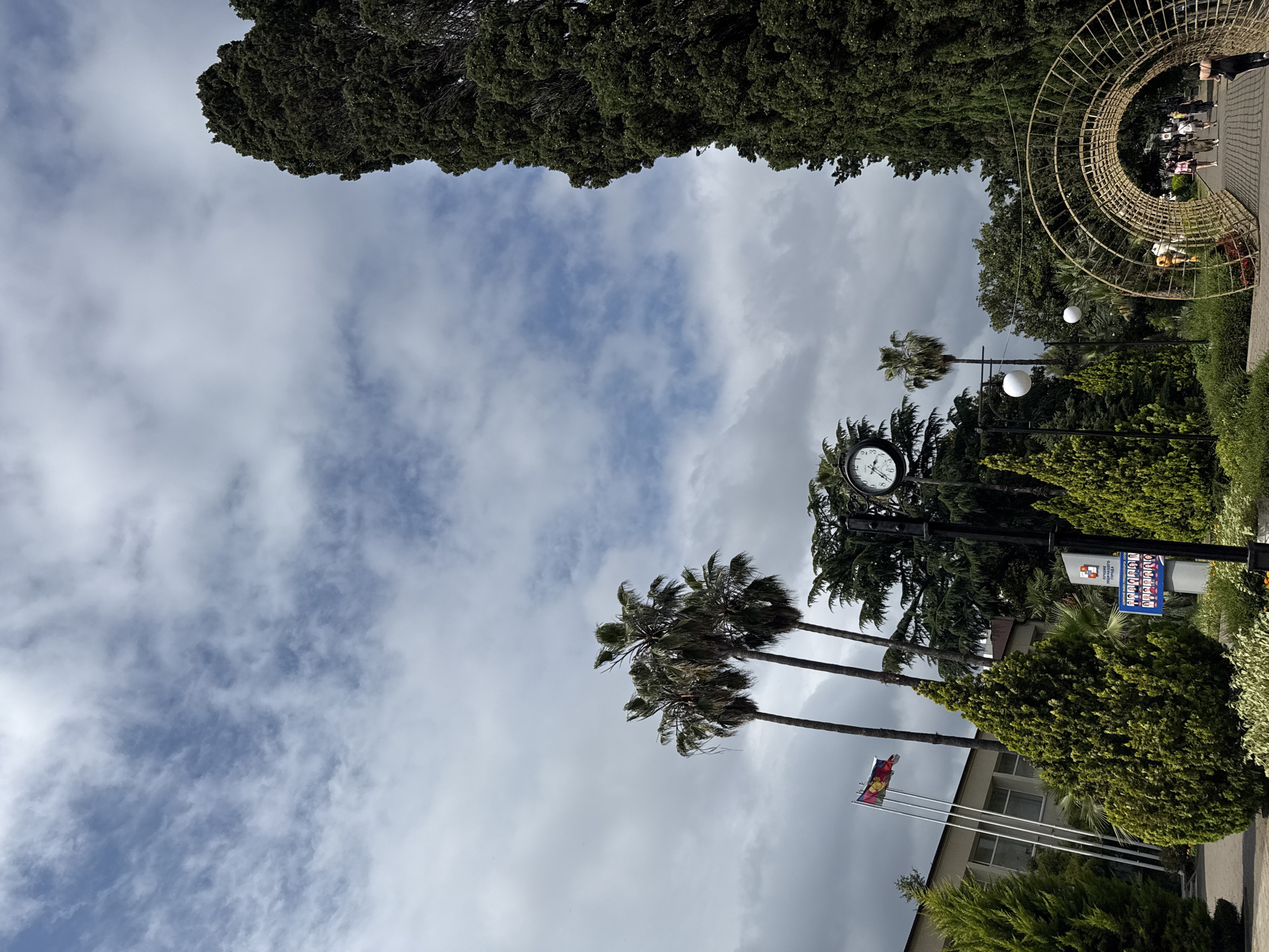 The image shows a park area in Sochi, Russia. The sky is partially cloudy with patches of blue visible. On the left side of the image, there is a building with flags flying in front of it. Three palm trees stand tall behind the flags. In the foreground, there are manicured evergreen bushes. A large clock is mounted on a pole in the middle of the scene. To the right, a tall, dark green tree dominates the frame. A pathway curves through the park, leading to a decorative archway. Several people are walking on the pathway under the archway. The overall scene depicts a public park with lush vegetation and architectural elements, under a partly cloudy sky.