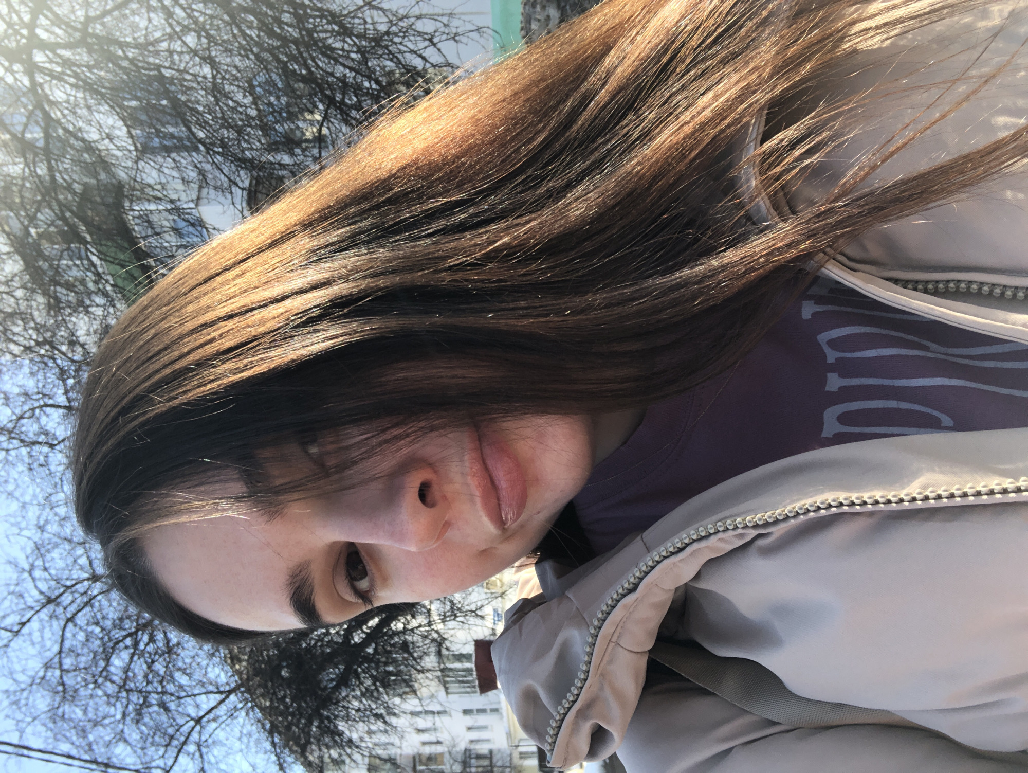 A young woman poses for a selfie outdoors, seemingly in the city of Chaikovsky, Russia, judging by the urban background visible in the lower portion of the image. The foreground is dominated by the woman's face, long brown hair, a beige jacket, and a glimpse of a purple t-shirt. Above her hair are bare tree branches against a light blue sky, possibly indicating late winter or early spring. The background building architecture suggests an urban setting.