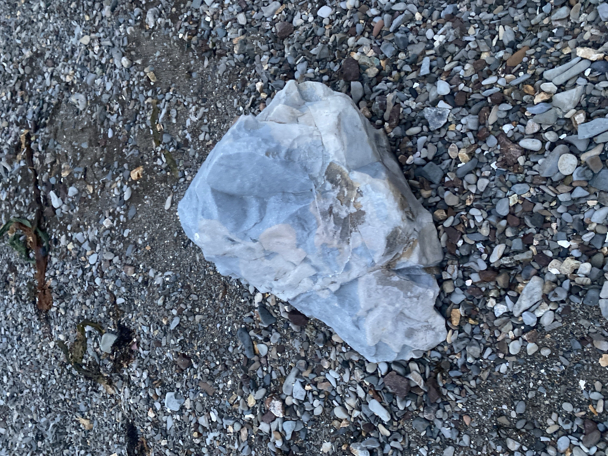The image shows a large, light-colored rock with rough, angular surfaces resting on a bed of small pebbles and stones. The ground is composed of a mixture of grey, brown, and some lighter-colored stones, with patches of darker, damp sand or soil visible. There are also some pieces of dark, stringy material, possibly seaweed or debris, scattered amongst the pebbles, particularly in the upper left portion of the image. The lighting suggests an overcast day, casting soft shadows. This scene is likely a beach or shoreline in the city of Korsakov, Russia, given the abundance of pebbles and the general appearance of coastal terrain. There are no people or animals visible in the image.