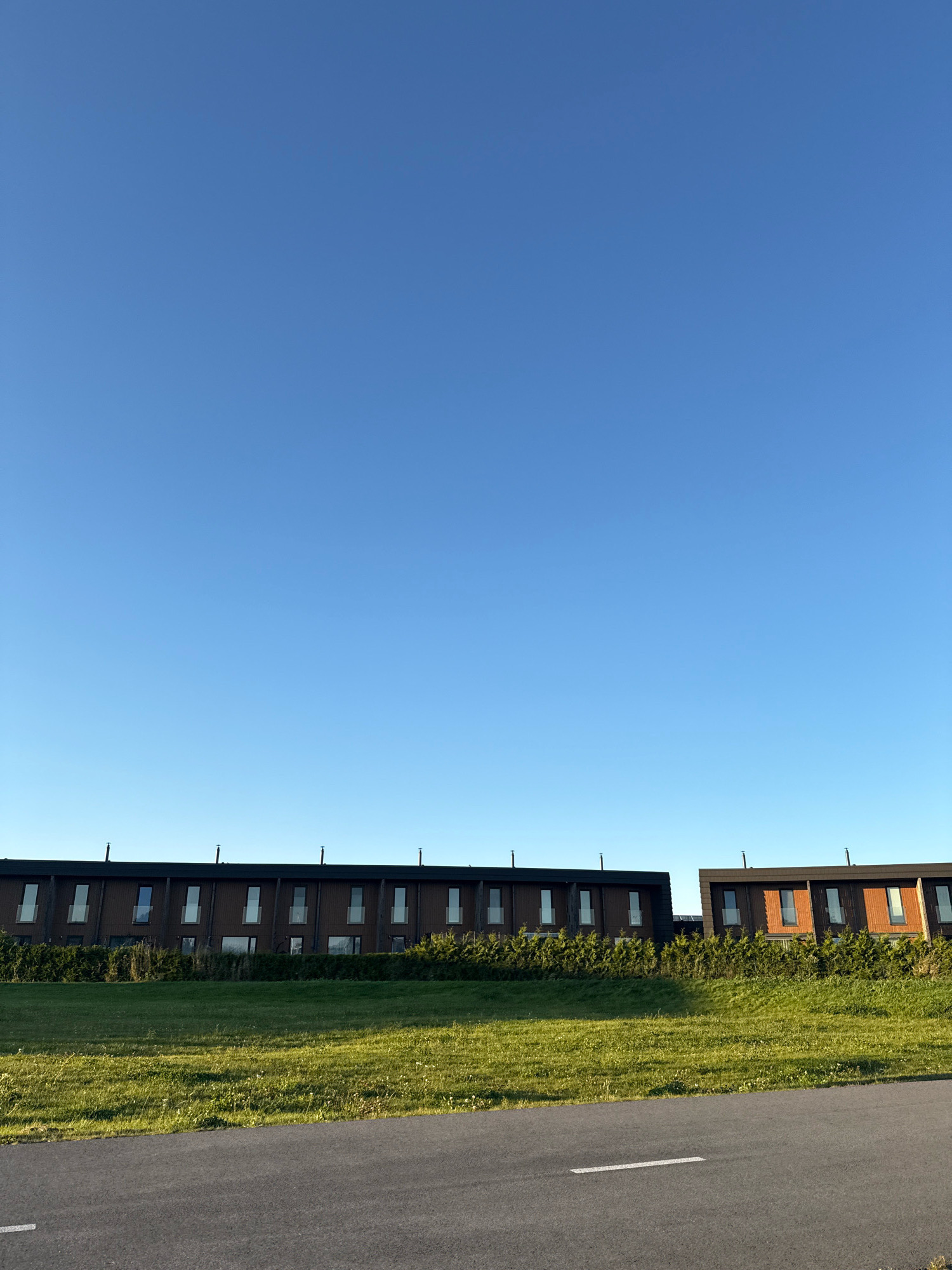 The image shows two modern buildings with dark brick facades and orange wooden accents, situated in Järveküla, Estonia. The buildings are rectangular and appear to be townhouses, each with multiple tall, narrow windows. They are separated by a small gap. The buildings are set against a backdrop of a clear, bright blue sky. In front of the buildings is a line of manicured green hedge. Below the hedge is a grassy slope that transitions into a paved road in the foreground. The road has a white line marking. The scene is bathed in the warm light of what appears to be late afternoon or early morning sun, casting shadows. There are no people visible in the image.
