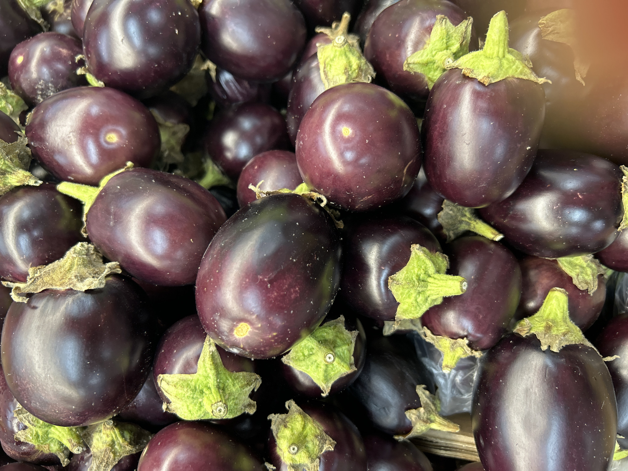 The image shows a close-up, overhead view of a pile of eggplants. The eggplants are dark purple and have green stems. They are tightly packed together, with some eggplants partially obscuring others. The lighting is bright, and there are some reflections on the surfaces of the eggplants. The scene appears to be in a market or grocery store. There is no indication of the city of Detmold, Germany, in the image or the provided context.