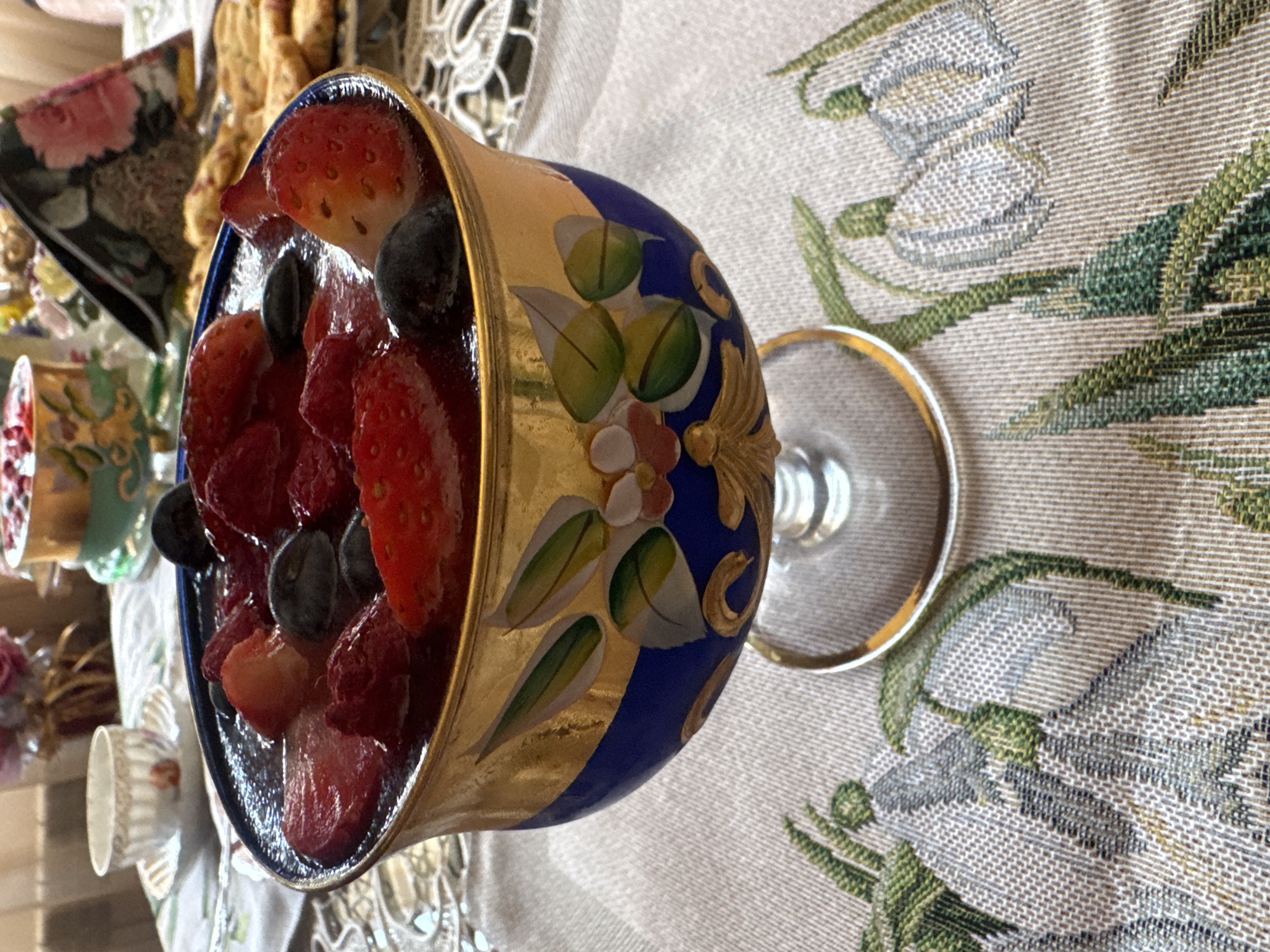 A close-up shot features a footed bowl filled with fruit, primarily strawberries and blueberries, sitting on a patterned tablecloth. The bowl is ornate, with a dark blue base, gold accents, and hand-painted floral designs. The fruit appears fresh and glistening, possibly glazed. The tablecloth has a neutral, linen-like texture with a floral pattern featuring grey and green hues. The background suggests an indoor setting, possibly a home or a cafe, with additional tableware visible on the left side of the frame. Given the nature of the image, the location in Plovdiv, Bulgaria cannot be determined. There is no information about people, actions, or interactions within the image.
