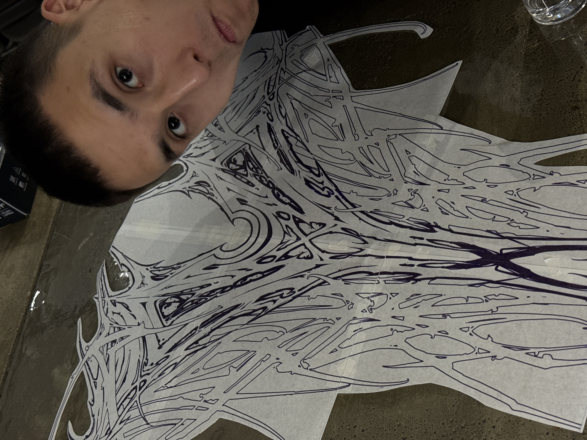 A selfie-style, high-angle shot depicts a person with short, dark hair and light skin looking at the camera. A detailed drawing, featuring intricate, swirling lines, is displayed on a piece of paper. The setting appears to be indoors, on a dark surface or table. The lighting is somewhat dim. There is a glass of water in the upper right corner and a dark object is visible on the bottom left corner. The location is unknown.
