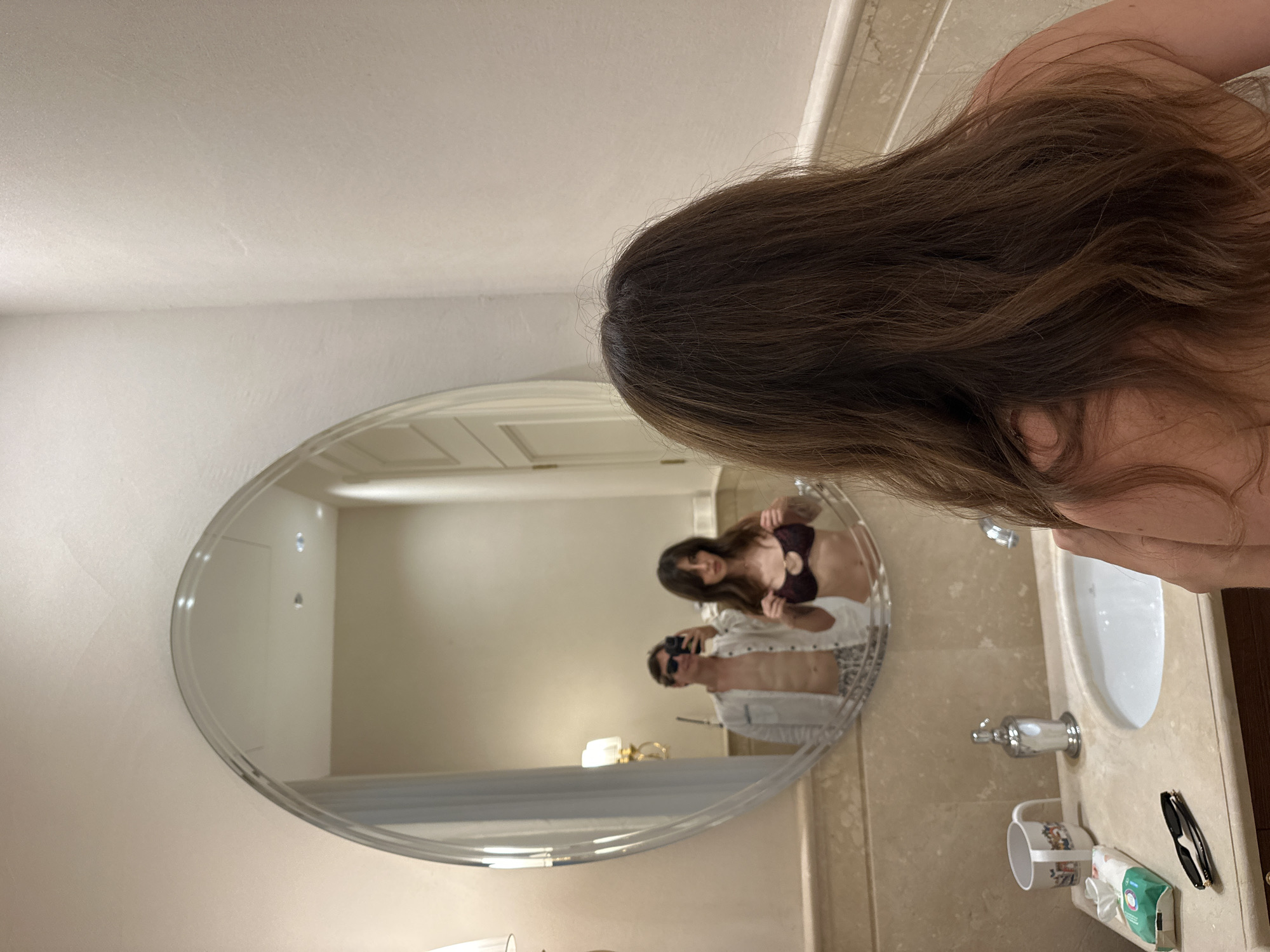 The image depicts a young woman with long, brown hair, seen from behind, standing in front of a bathroom mirror. In the mirror's reflection, a man wearing sunglasses, an open white shirt, and patterned shorts is visible. He appears to be holding a phone, taking a selfie. Beside him in the reflection, a woman, presumably the same woman as in the foreground, is adjusting a dark-colored bikini top. The bathroom features a marble countertop with a sink, a soap dispenser, a mug, a pack of wet wipes, and a pair of sunglasses. The overall environment suggests a residential bathroom, potentially in the city of Savyon, Israel.