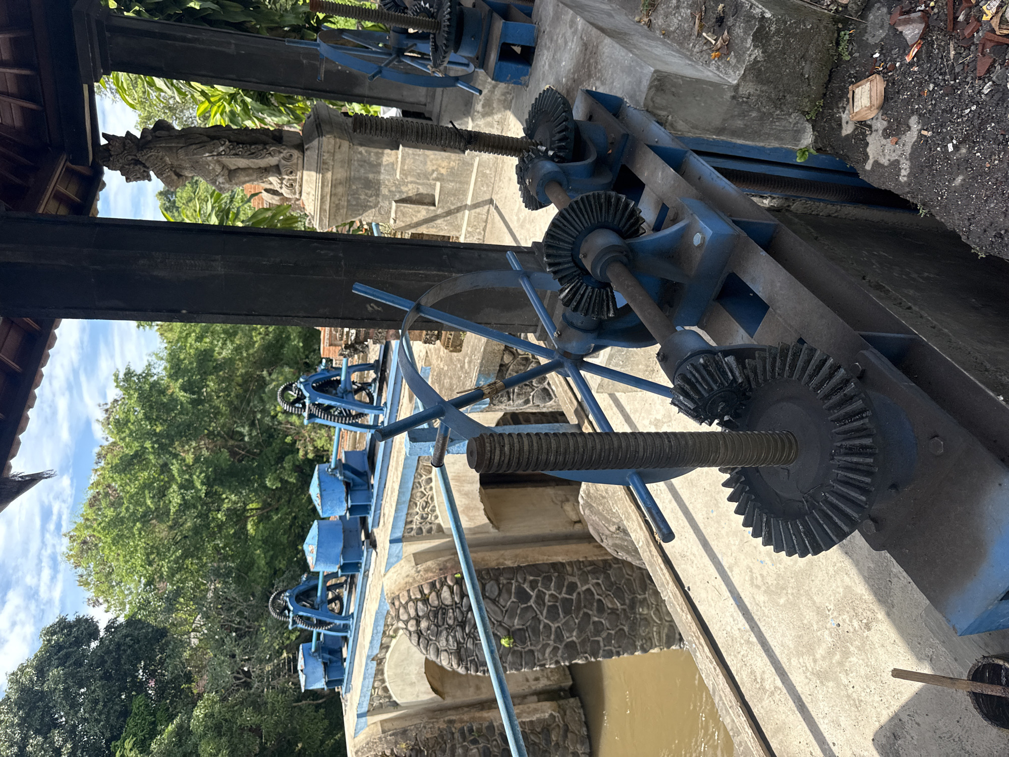 This image shows a system of blue metal gears and structures built over a waterway. The gears appear to be part of a mechanism used to control water flow. The structure is made of concrete and stone. The water is brown. In the background, there are trees and a cloudy sky. A statue can be seen on top of a stone pillar. This is likely a water management system, possibly an irrigation dam, located in Denpasar, Indonesia.