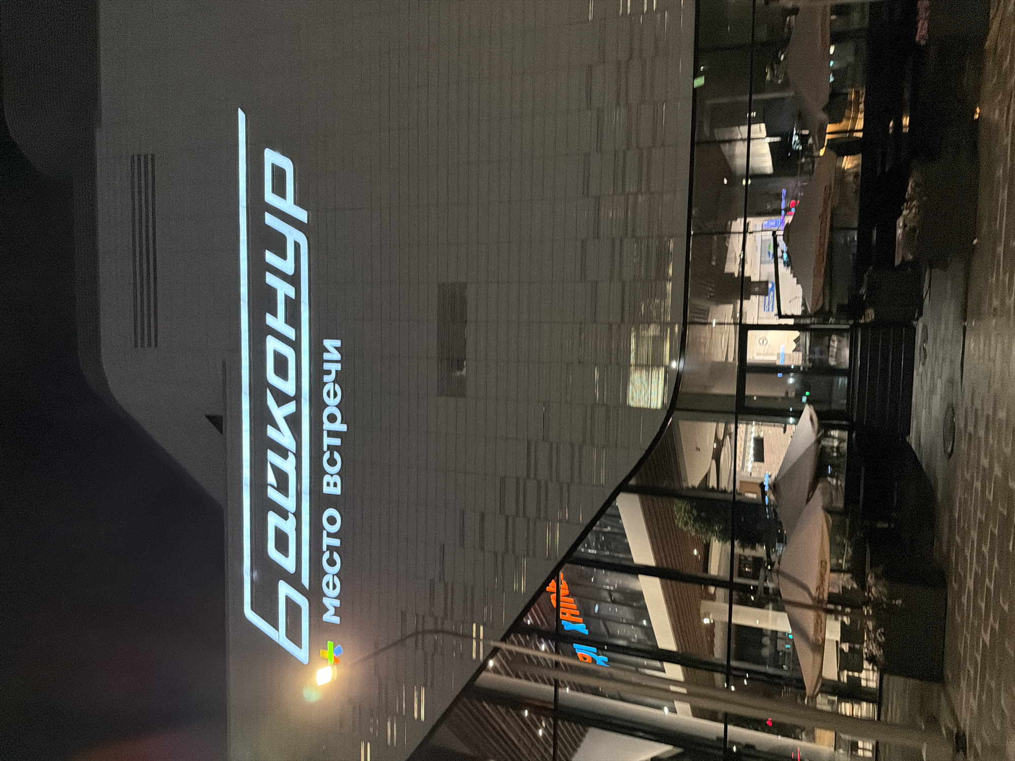 The image shows the exterior of a building at night, with a large sign reading "БАЛКОНЦ" in Russian, and below it, "место встречи", which translates to "meeting place". The building's facade is primarily white with a grid-like pattern and large glass windows. Reflections on the glass show interior details such as white umbrellas and interior spaces. The surroundings are dimly lit, suggesting an urban environment, possibly a business or commercial district in Moscow, Russia. There are hints of other signage and architectural details reflected in the windows, but specific individuals or activities are not apparent. The scene appears to be at night with artificial lighting illuminating the sign and surrounding areas.