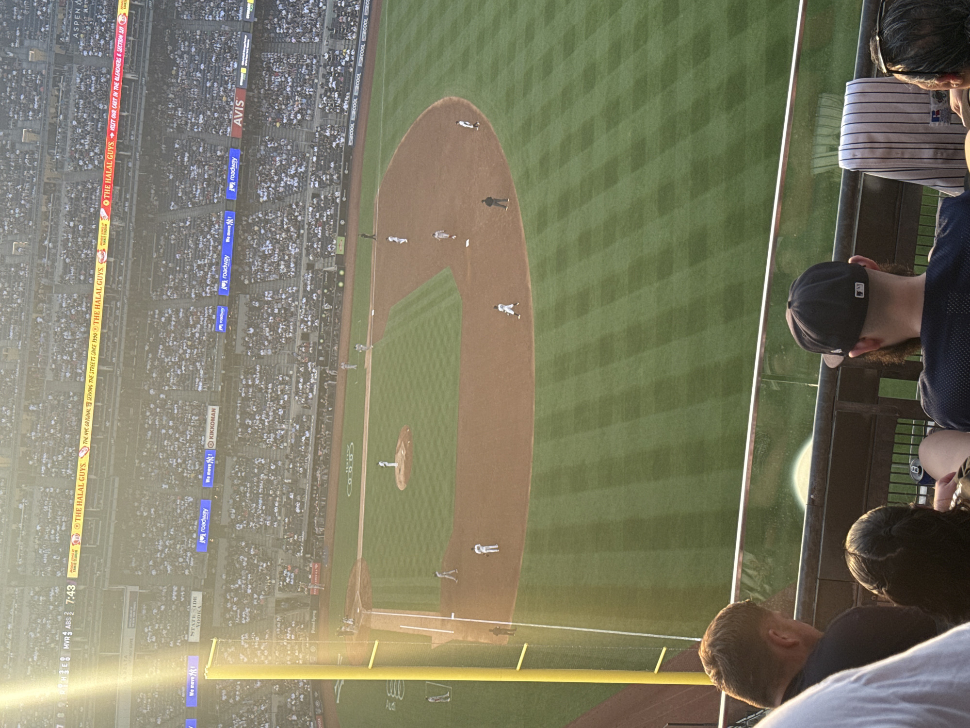 This high-angle image depicts a baseball game in progress at Yankee Stadium in The Bronx, New York City, United States of America, under late afternoon or early evening sunlight causing significant lens flare on the left. In the foreground, several spectators are visible from behind, observing the field from an upper deck. One individual on the far right wears a navy baseball cap with a white MLB logo and a dark sleeveless shirt, with a striped New York Yankees jersey draped over the railing in front of them. A glass barrier and dark railing separate the spectators from the playing field.
The midground features the baseball diamond with a game underway. A pitcher is on the mound, a batter is at home plate, and a catcher is positioned behind. Runners are visible on second base and near first base, with a coach or umpire near third base. The field exhibits distinct green grass patterns in the outfield and dirt infield areas. A tall yellow foul pole stands prominently on the left side of the field.
The background shows multiple tiers of stadium seating filled with a large crowd of spectators, predominantly in light-colored attire. Various illuminated advertising banners and screens are integrated into the stadium architecture. Prominent advertisements include "THE HALAL GUYS" (identified as "PROUD PARTNER OF YANKEE STADIUM" and "THE NYC ORIGINAL SERVING THE STREETS SINCE 1990"), "STATE SIDE VODKA," "roadway" (multiple instances), "We move MVR!", "KIKKOMAN," and "AVIS." A scoreboard or screen in the upper left displays "MVR 3 ABS 2" and "7:43."