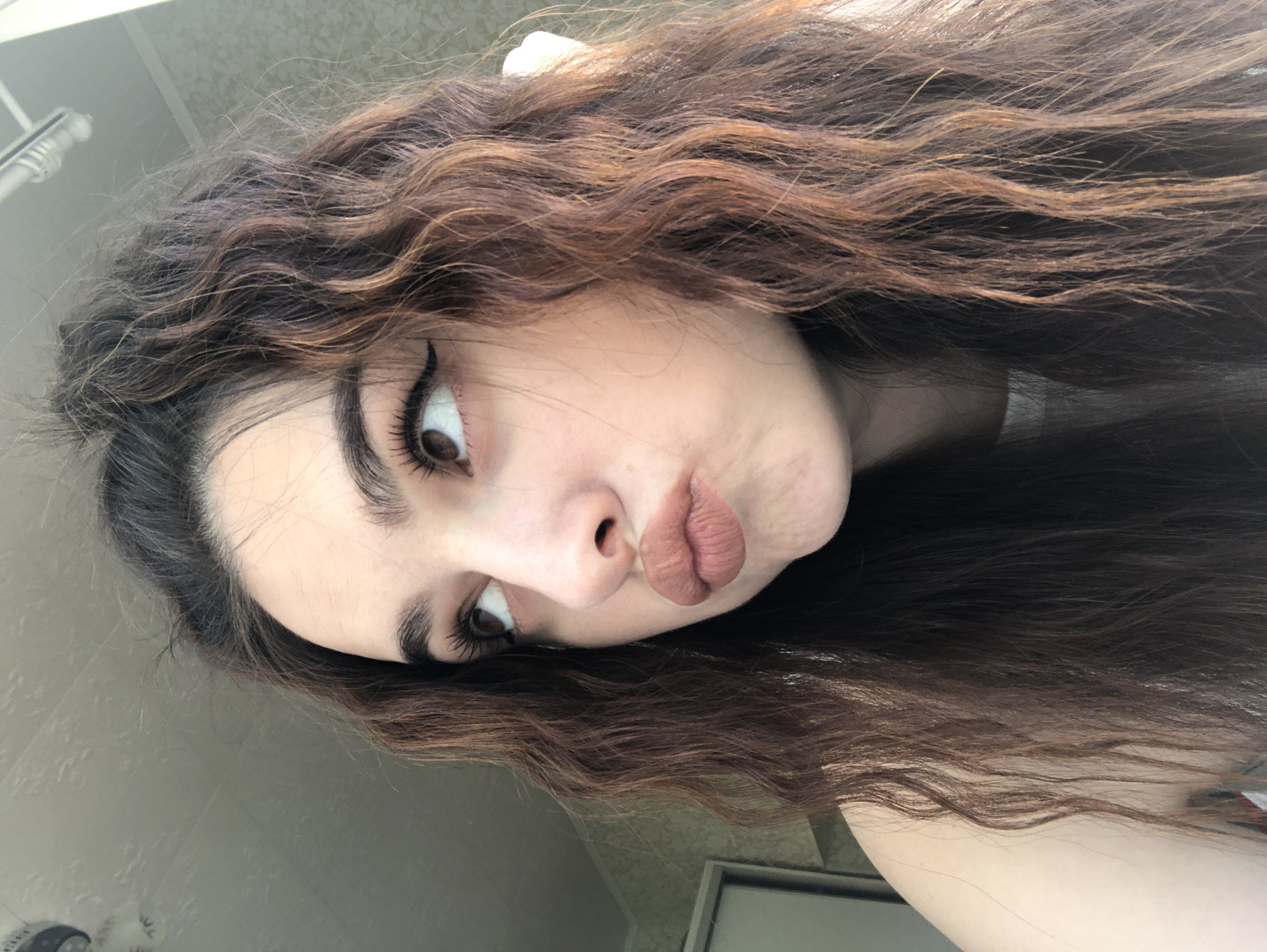 A woman with long, wavy brown hair and distinct eyeliner is making a "duck face" pose. The background appears to be an interior setting, possibly a bathroom, with patterned wallpaper or tile visible in the upper left. The image is rotated. There is no information to indicate the location is Noviy, Russia.