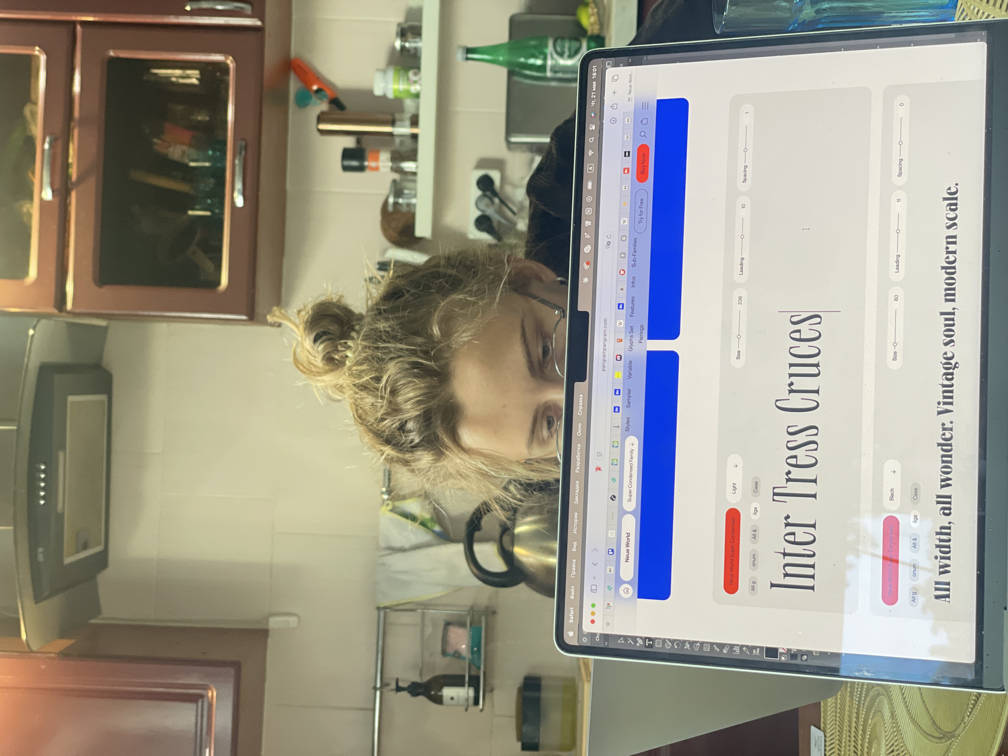 A person with light hair in a bun is looking at a laptop screen displaying a website with font customization options. The screen shows the words "Inter Tress Cruces" in a large font, along with sliders for size, leading, and spacing. The website, pangrampangram.com, appears to be a resource for font design. The location is an indoor space, likely a kitchen, with cabinets, shelves, and kitchen appliances visible in the background. The laptop is on a patterned surface. There is no indication that this is in Sestroretsk, Russia.