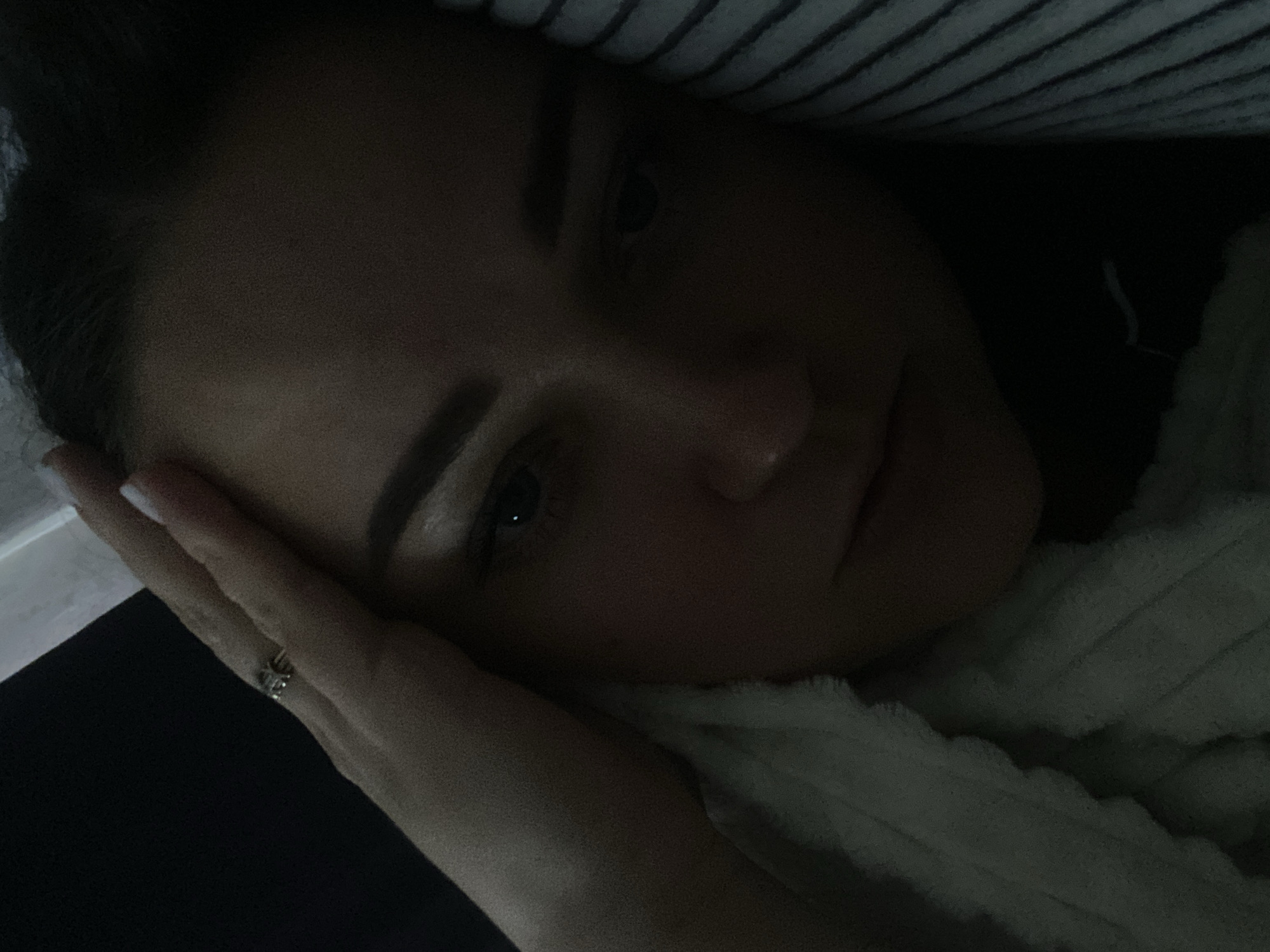 A close-up, low-light photograph captures a woman resting her head on her hand. A blanket or possibly a duvet is visible to the right of her face. The background features horizontal stripes. A ring is visible on her finger. The location is not identifiable from this image.