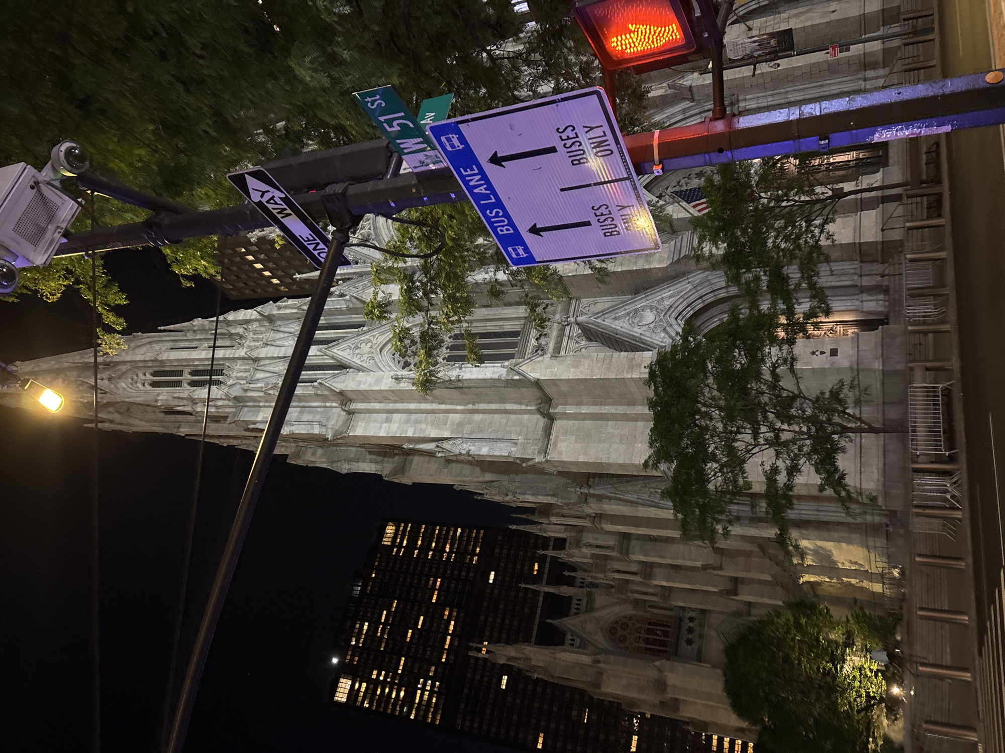 This vertical-format, nighttime photograph captures a street scene in New York City, featuring a prominent Gothic Revival cathedral and modern urban elements. The majestic stone cathedral, with its intricate spires, arched windows, and detailed façade, rises dramatically against the dark sky. To its right, a contemporary high-rise building with numerous illuminated windows suggests offices or residences.
In the foreground, a traffic light glows red, and several street signs are mounted on a pole. A sign with a white arrow clearly indicates "ONE WAY." Below it, a larger blue-and-white sign designates a "BUS LANE" with two arrows pointing forward, and the text "BUSES ONLY" is visible twice. Above these, green street signs confirm the location as "W 51 ST" and "5 Av."
Security or traffic monitoring cameras are attached to a pole on the left, near some leafy green trees. A single streetlight casts a yellow glow, illuminating part of the scene. An American flag is visible, hanging near one of the cathedral's entrances. The image presents a blend of historic architecture and modern city infrastructure, characteristic of New York City at night, with no people or specific activities taking place.
