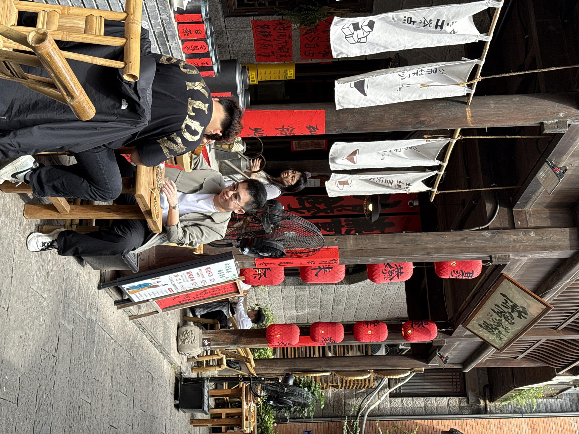 The image shows an outdoor scene in what appears to be a traditional setting, likely in the city of Yuzhong District, China, given the presence of Chinese text and architectural elements. Two men are sitting on bamboo chairs at a table. One man, wearing a black jacket with gold lettering, is leaning towards the other, who is wearing a gray jacket and glasses. They appear to be engaged in a conversation or transaction. The ground is paved with stone. Hanging above are a series of white flags with different black images and Chinese text on them, red lanterns, and signs with Chinese characters. Also visible are several other people in the background, including one lying on a bamboo chair. The scene includes elements such as a menu board, fans, and decorative objects that suggest a cafe or restaurant environment.