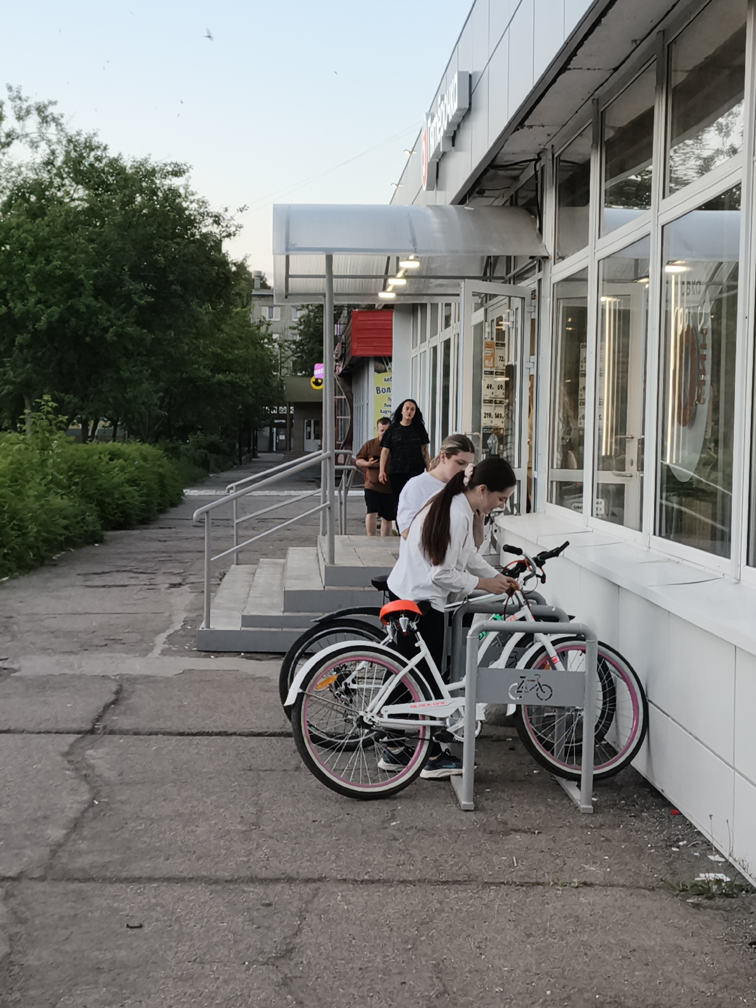 The image depicts an outdoor scene in front of a commercial building. In the foreground, there are several bicycles parked at a bike rack. Two young women are interacting with the bicycles, with one appearing to be unlocking or adjusting one of them. Behind them, the facade of the building features large windows and a canopy. Further back, on the sidewalk, a man and another woman are visible, walking or standing. The environment includes a paved walkway, green foliage, and a cloudy sky. The location is identified as Chaikovsky, Russia, with the commercial building likely being a local establishment.