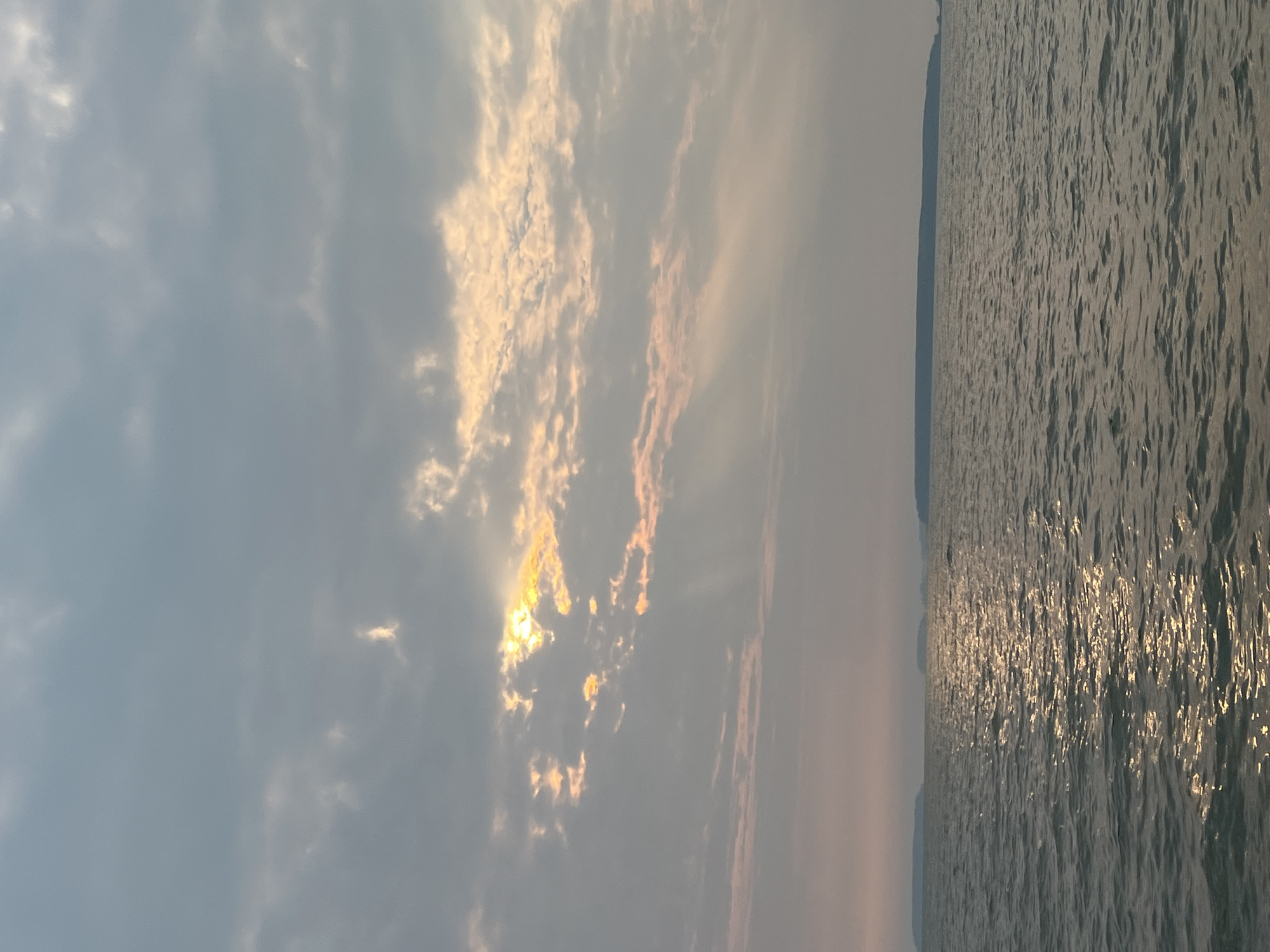 The image depicts a tranquil seascape with a cloudy sky and calm waters. In the distance, several islands or peninsulas are visible as dark silhouettes against the horizon. The sun is partially obscured by clouds, casting golden rays that illuminate a portion of the sky and reflect on the water's surface, creating a shimmering effect. The overall mood of the image is serene and peaceful. The location of Novopichugovo, Russia, is not discernible from the visual information present in the image.