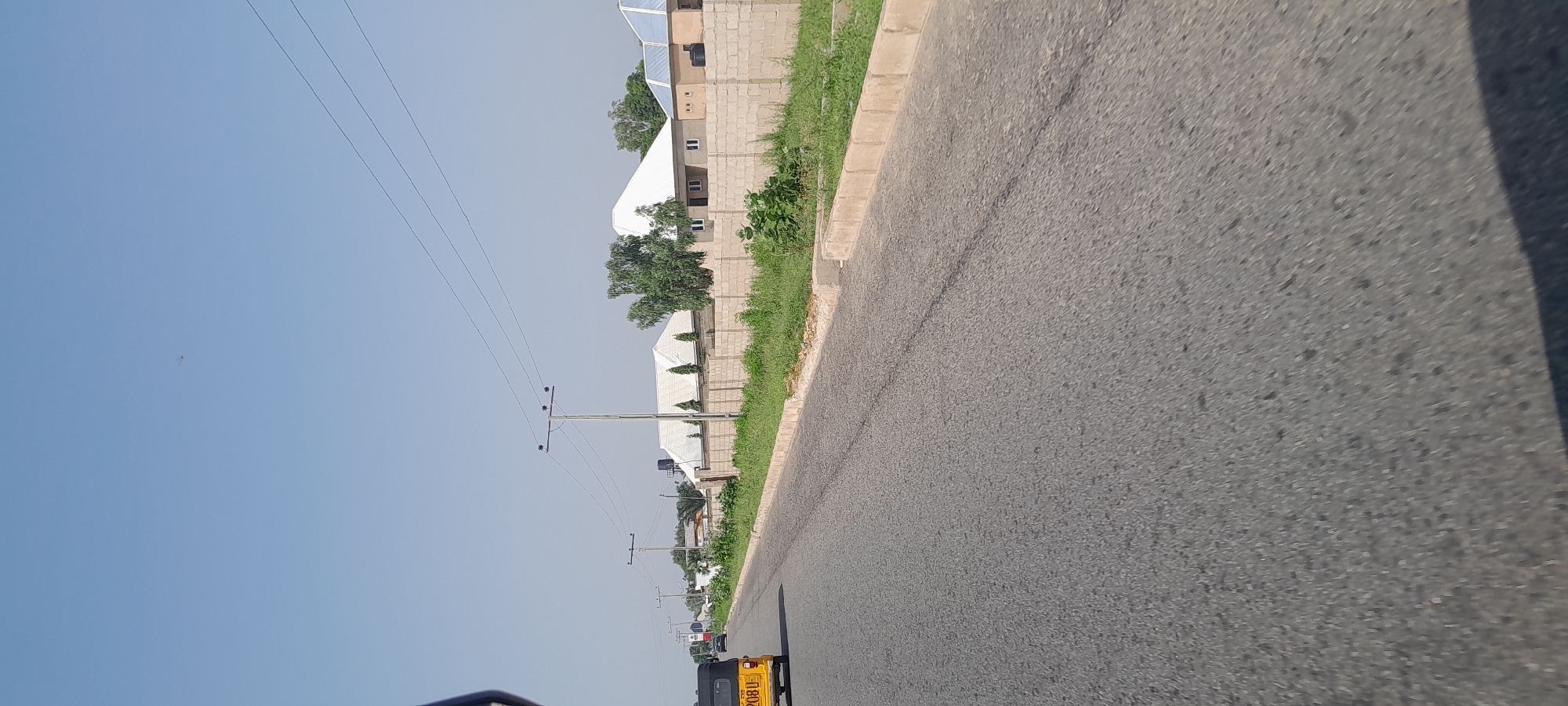 The image depicts a view of a paved road in Jalingo, Nigeria, viewed from a moving vehicle. The road surface is asphalt, showing some wear and tear with visible cracks and patches. To the left of the road, there is a grassy verge bordered by a light-colored concrete curb. Beyond the verge, a row of buildings and houses with white roofs and light-colored brick walls are visible. Some of these structures have solar panels on their roofs. Trees and other greenery are interspersed among the buildings.
Power lines supported by utility poles run parallel to the road, indicating an urban or semi-urban setting. In the distance, several vehicles are seen on the road, including a yellow trishaw-like vehicle in the foreground, suggesting ongoing traffic. The sky is clear and bright blue, indicating it is daytime, likely late morning or afternoon given the lighting. There are no discernible emotions conveyed by the scene; it is a straightforward depiction of a street view.
Visible text on the yellow vehicle reads "208U". The overall scene suggests a typical day in a Nigerian town or city, with infrastructure and everyday life elements visible. The low angle of the shot implies it was taken from a low-lying vehicle or possibly a bicycle.
