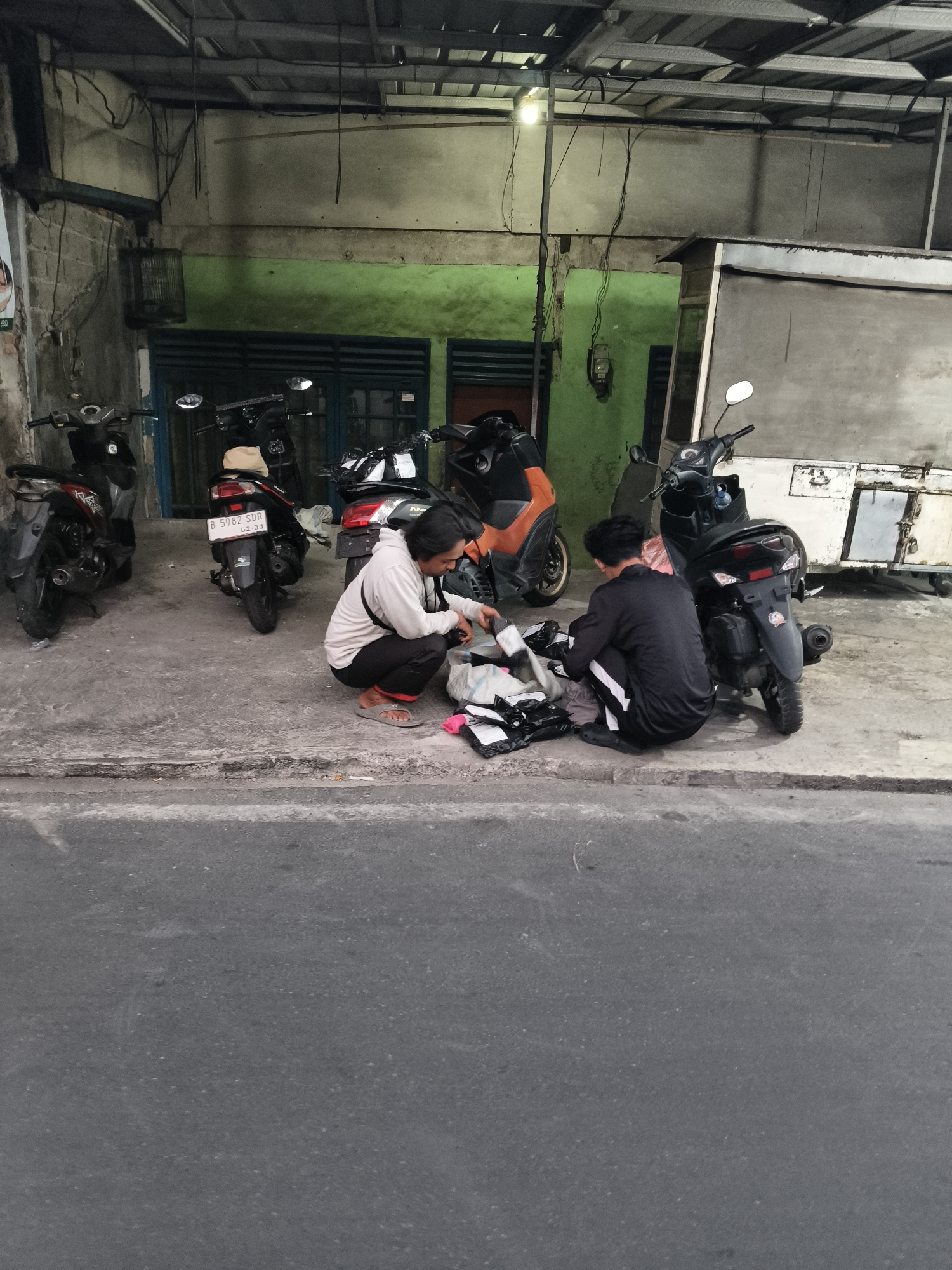 The image depicts a street scene in South Tangerang, Indonesia, where two individuals are crouched down sorting packages. The setting appears to be an alleyway or side street adjacent to a building with a green wall and a corrugated metal roof. Several motorcycles are parked in the background.
One individual, wearing a light-colored hoodie and dark pants, is on the left, facing towards the packages. The other individual, dressed in a black tracksuit with white stripes, is on the right, also engaged with the packages. Both are focused on the task at hand, with various packages, some black and some white, spread out on the ground between them.
The environment is somewhat utilitarian, with visible electrical wiring overhead and what appears to be a cage hanging from the wall. The ground is paved concrete, and the street in the foreground is asphalt. The lighting suggests it might be dusk or an overcast day. The overall impression is one of everyday activity, possibly related to delivery or retail.