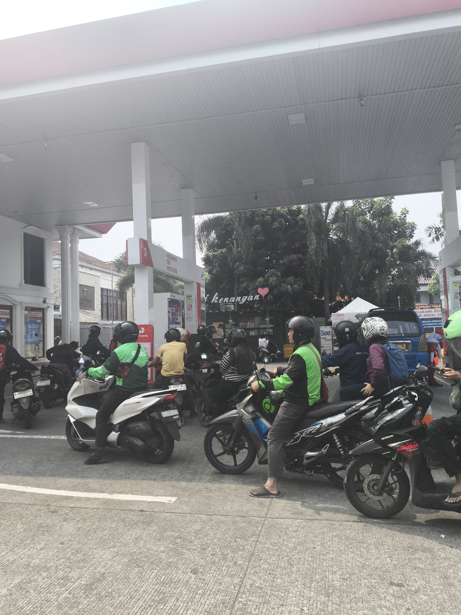 The image captures a scene at a Pertamina gas station in Depok, Indonesia, on a bright, possibly overcast day. A long line of motorcycles is queued up at the pumps, indicating a busy period. Many of the riders are wearing helmets and some are wearing bright green jackets, suggesting they might be delivery drivers for a service like Gojek.
The gas station canopy, supported by white pillars, extends over the pump area. A Pertamina sign with the number "3" and text indicating "Pertamina Dex" and "Pertalite" is visible on one of the pumps. Behind the motorcycles, there are trees and buildings, providing context to the urban environment of Depok. The ground is paved with concrete, and a white line marks a lane or boundary.
In the foreground, the focus is on the motorcycles and their riders. One rider in a green jacket is prominently featured, as are other individuals on their bikes. The overall atmosphere suggests a routine, everyday activity of refueling vehicles in a bustling city.