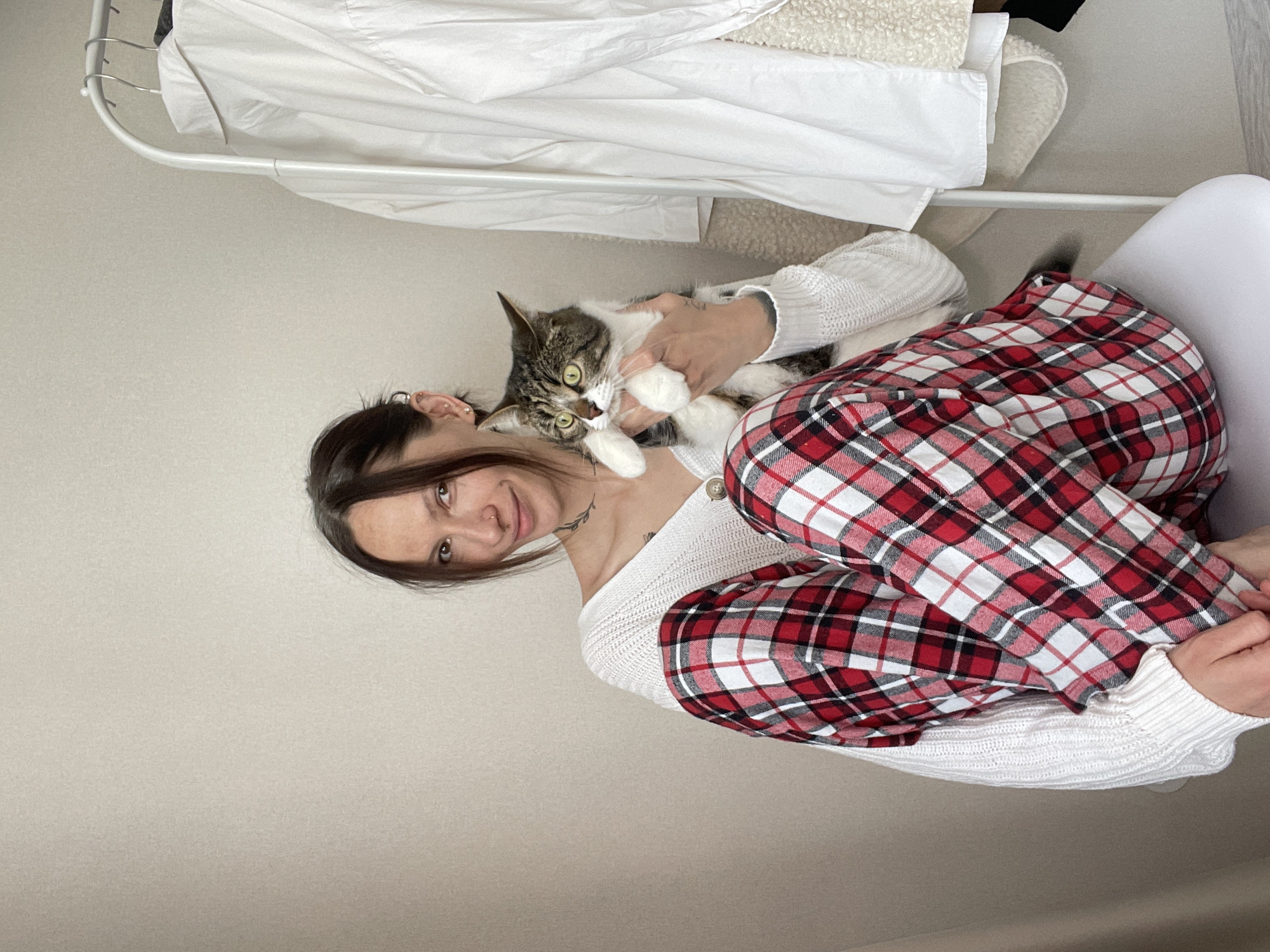 A woman is sitting in a white chair and holding a tabby cat in her arms. She is wearing a white sweater and red plaid pajama pants. Behind her is a clothing rack with white shirts and a cream-colored jacket. The room has light gray walls. The location cannot be determined to be in the city of Pervomaysky, Russia.