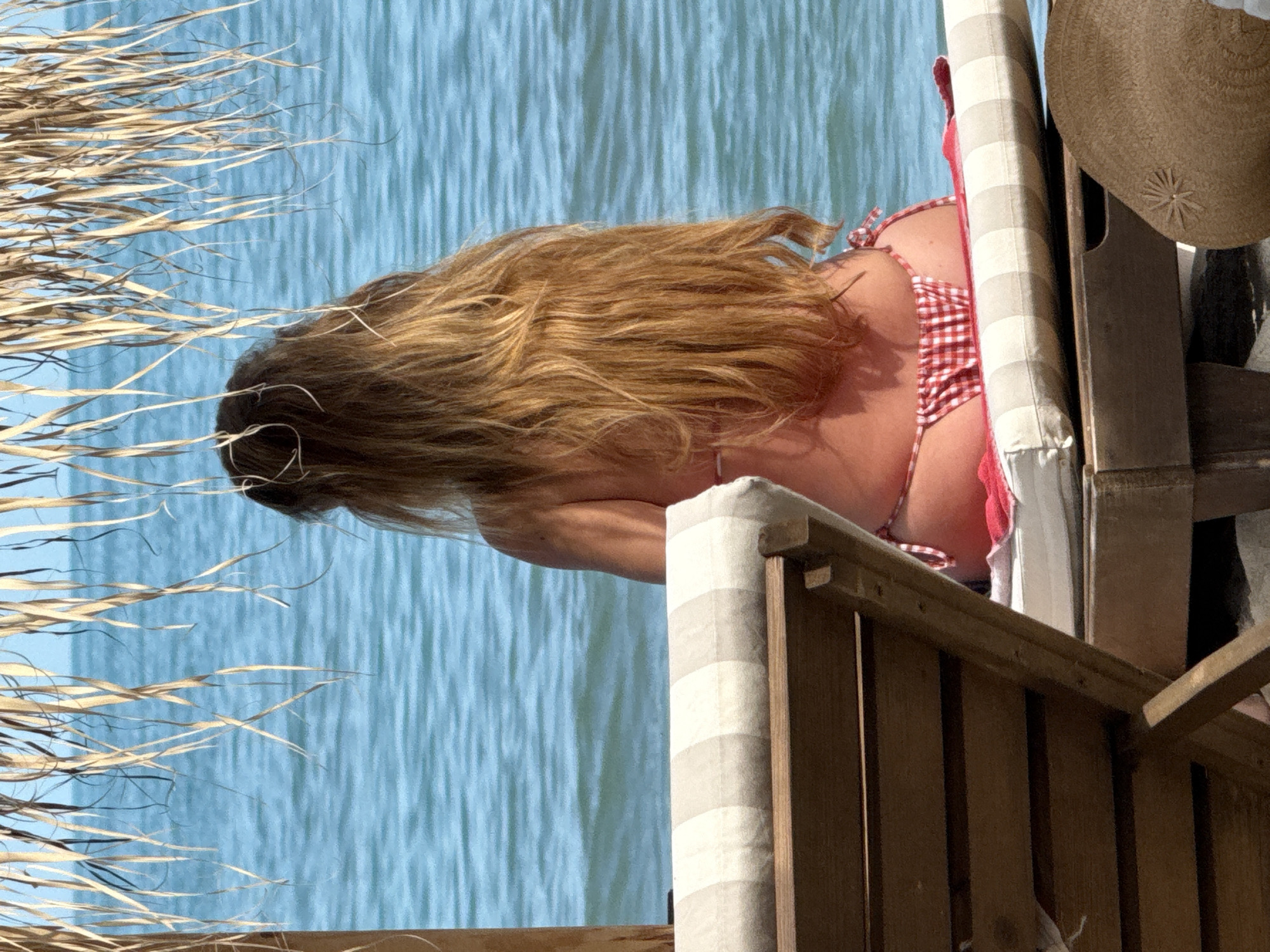 A person is photographed from behind, seated on a wooden sun lounger at a beach in Kranevo, Bulgaria, facing a calm, blue sea with minimal ripples. The individual possesses long, light brown hair and is wearing a red and white gingham-patterned bikini bottom. A red towel is placed beneath the person on the sun lounger's beige and white striped cushion. Above, dried straw or reeds from a beach umbrella or structure provide overhead cover. In the lower right foreground, a light brown straw hat with a central decorative element is visible. The scene is illuminated by natural daylight.