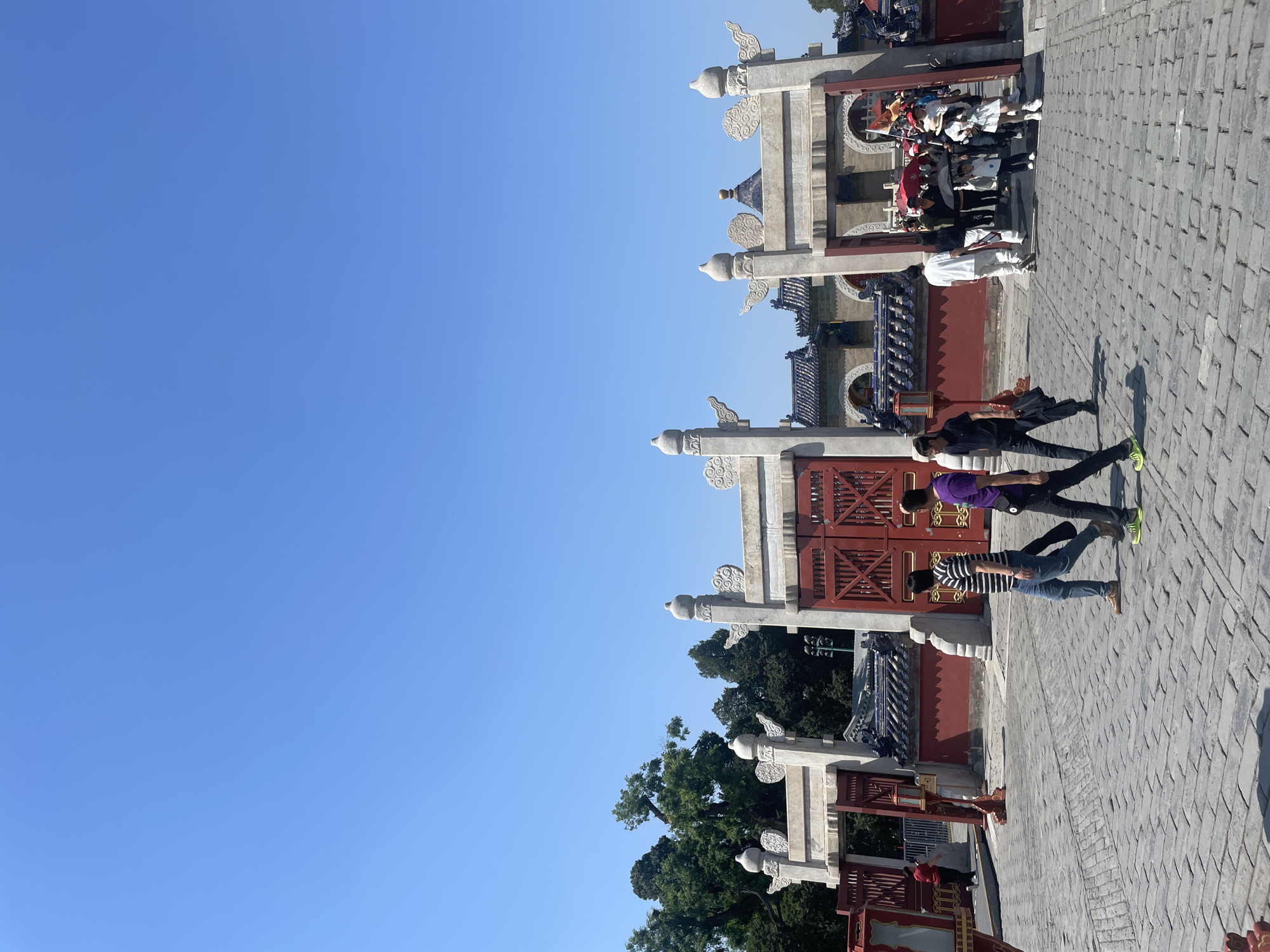 The image shows a view of a traditional Chinese architectural structure, possibly a gate or entrance, located in the Dongcheng District of China. The structure is made of stone and wood, with intricate carvings and a red-painted finish on the wooden components. Two people are captured in the scene, casting shadows on the stone-paved ground. The sky is clear and blue, indicating a sunny day. Green trees can be seen in the background, suggesting a park or garden setting. People are walking through the gate. The structure is likely part of a historic site or temple complex in the Dongcheng District.