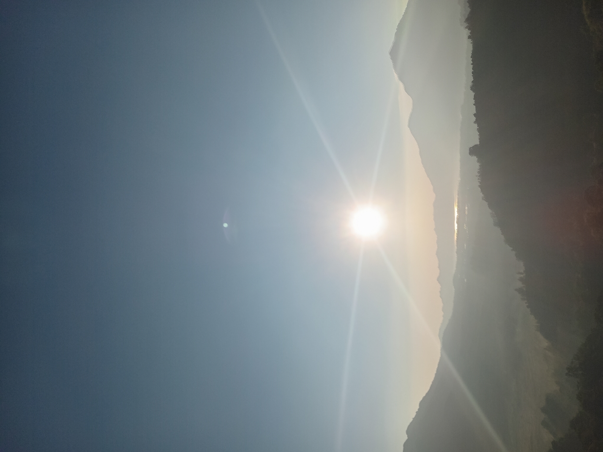 The image captures a panoramic view of the Kintamani region in Bali, Indonesia, during sunrise. The sun is a bright, radiant orb in the sky, casting a warm glow and creating lens flares that streak across the frame. The foreground is dominated by rolling hills and valleys, shrouded in a soft morning mist that lends a hazy, ethereal quality to the landscape. Silhouetted against the brightening sky are the majestic outlines of mountains, some with jagged peaks, others more rounded. In the distance, a body of water, likely Lake Batur, is visible, reflecting the nascent light of day. The overall atmosphere is serene and tranquil, evoking the peaceful beauty of a Balinese dawn. There are no people or animals visible in the image.