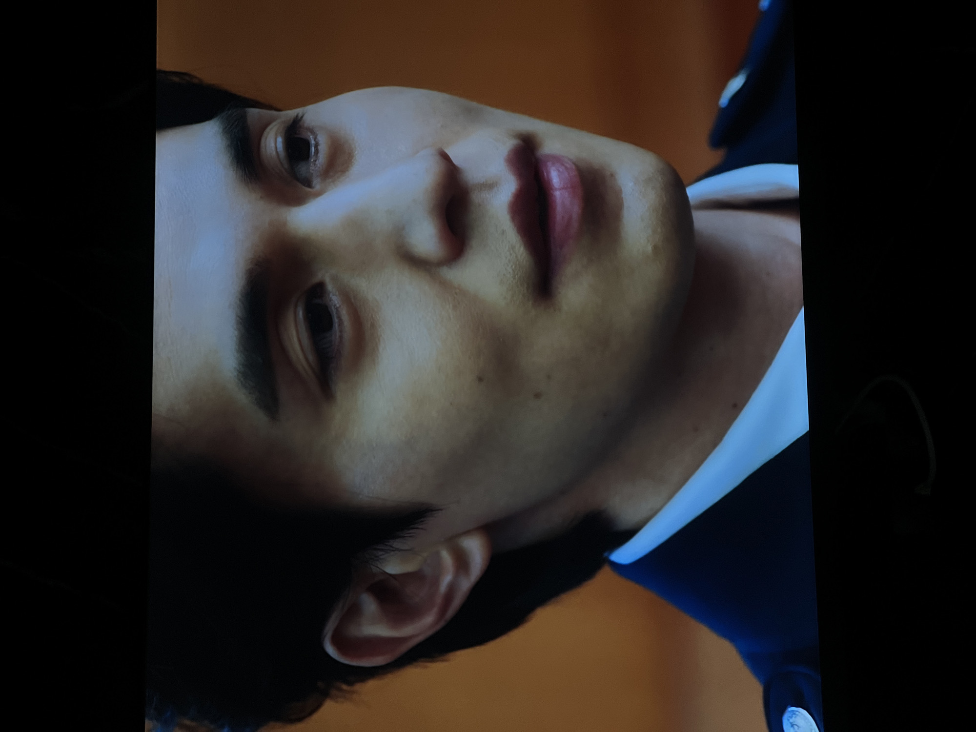 A close-up, slightly rotated, image captures the face of a man with dark hair. His eyes are open, and his expression appears neutral. He has visible facial features, including eyebrows, nose, lips, and an ear. The man is wearing what appears to be a collared shirt or jacket with visible buttons. The background is blurred and out of focus, presenting a warm, muted color. There is no discernible indication of the city of Vologda, Russia, or any other geographical location.