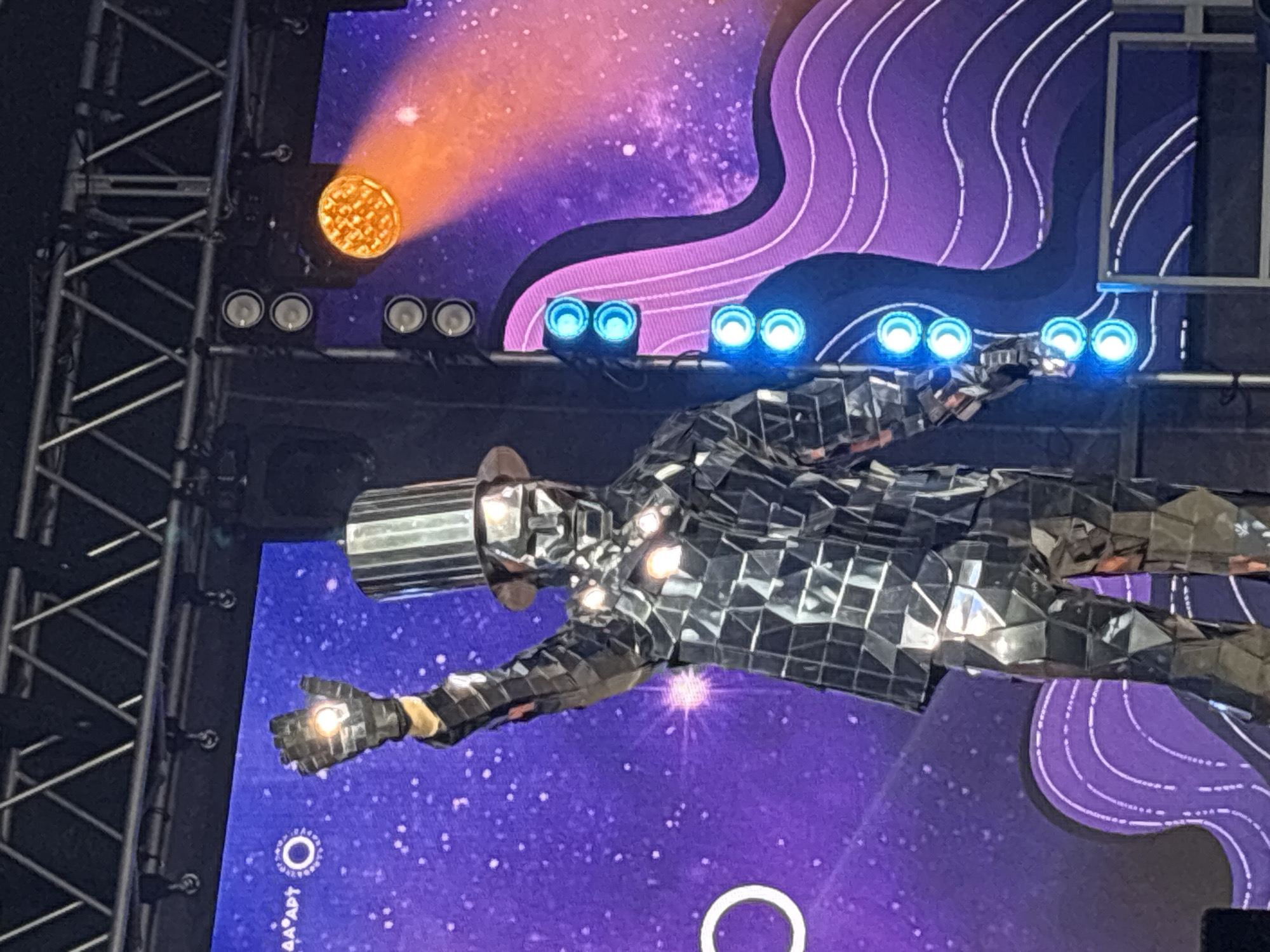 A performer, clad in a mirrored suit and a tall, cylindrical, silver hat, stands center stage with their right arm raised. The performer's face is obscured by a metallic mask. The suit is covered in numerous small, square, reflective pieces, creating a dazzling effect. The performer is wearing a black glove on their raised hand, which appears to have a light or reflective element on the palm. The background consists of a large LED screen displaying a purple starry sky with a bright orange light streak on the right side. To the left of the performer, a large white circle is visible on the screen. Stage lighting, including multiple blue lights arranged vertically on the right side of the frame and an orange spotlight above, illuminates the scene. The stage structure, with metal trusses and lighting equipment, is visible at the top of the image. The overall setting appears to be an indoor venue or outdoor stage at night.