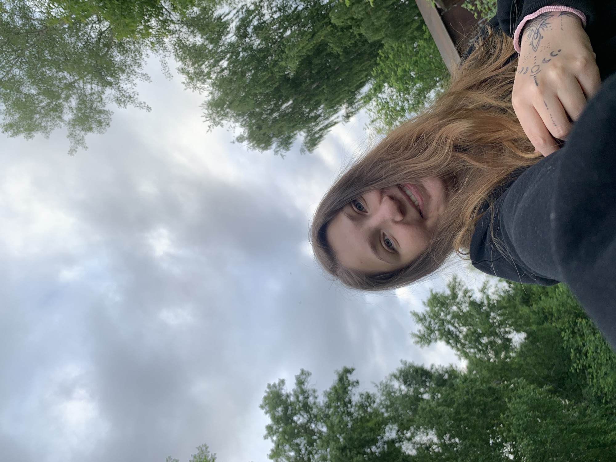 A young woman with long, wavy brown hair smiles at the camera in a selfie taken outdoors. She is wearing a black hoodie. Her right hand, adorned with intricate black tattoos and a pink bracelet, is visible in the foreground. The background consists of lush green trees and a cloudy sky. The scene appears to be a park or backyard, possibly in the city of Chaikovsky, Russia, given the natural setting.